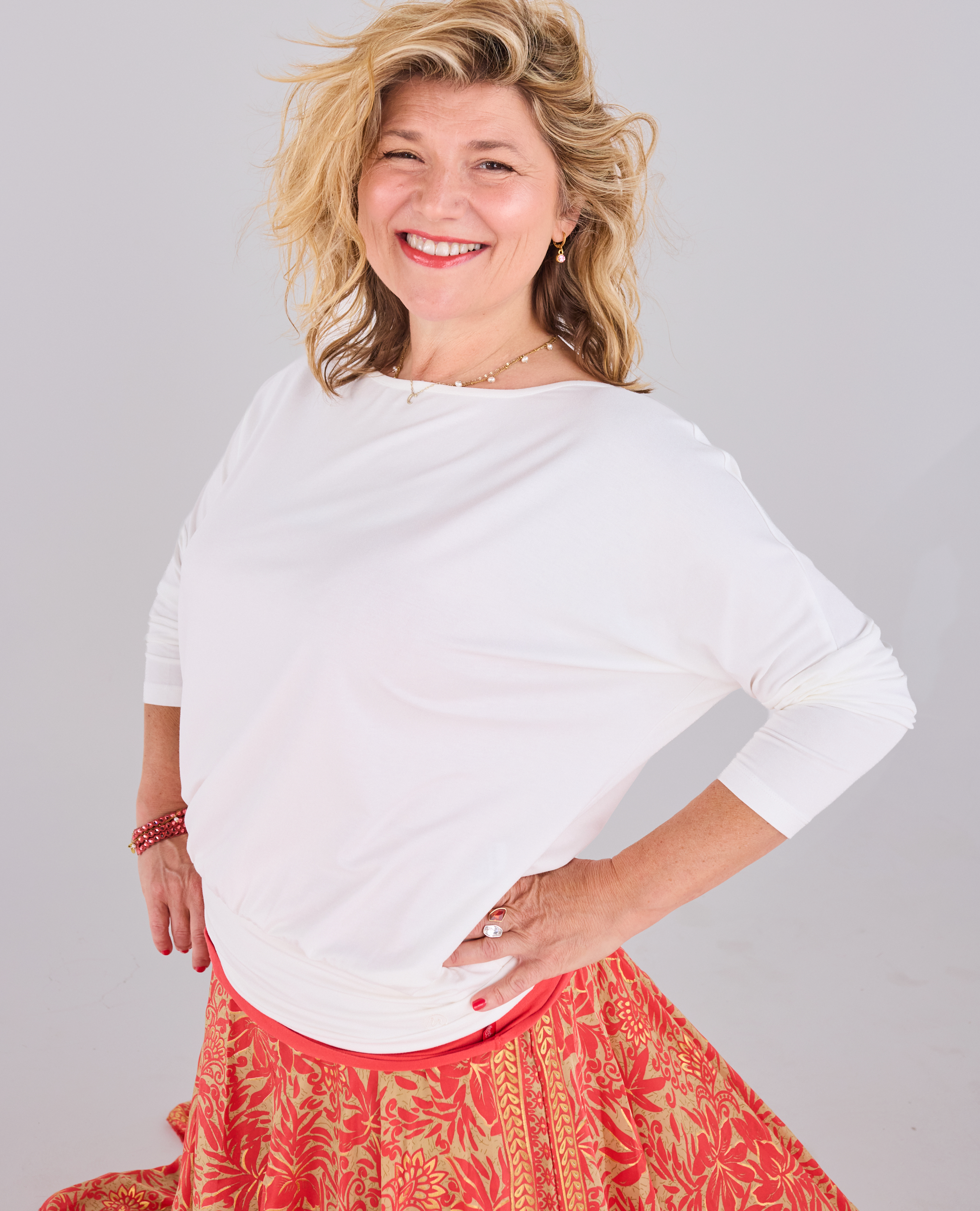 A smiling woman with blonde hair wearing a Melyr white Aimee boat-neck long-sleeve top and a red and orange patterned skirt, posing against a light grey studio background.