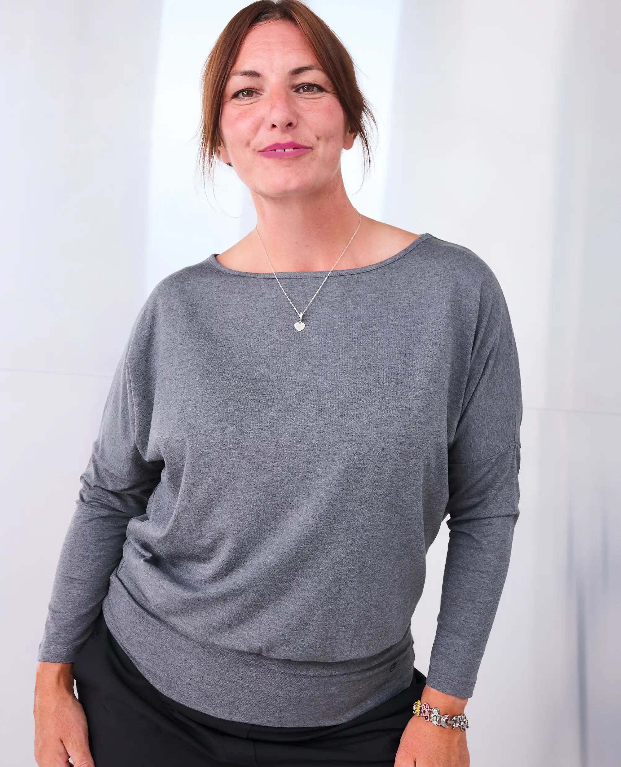 A woman wearing a Melyr Aimee grey long-sleeve boat-neck top and black bottoms, standing against a silver background.