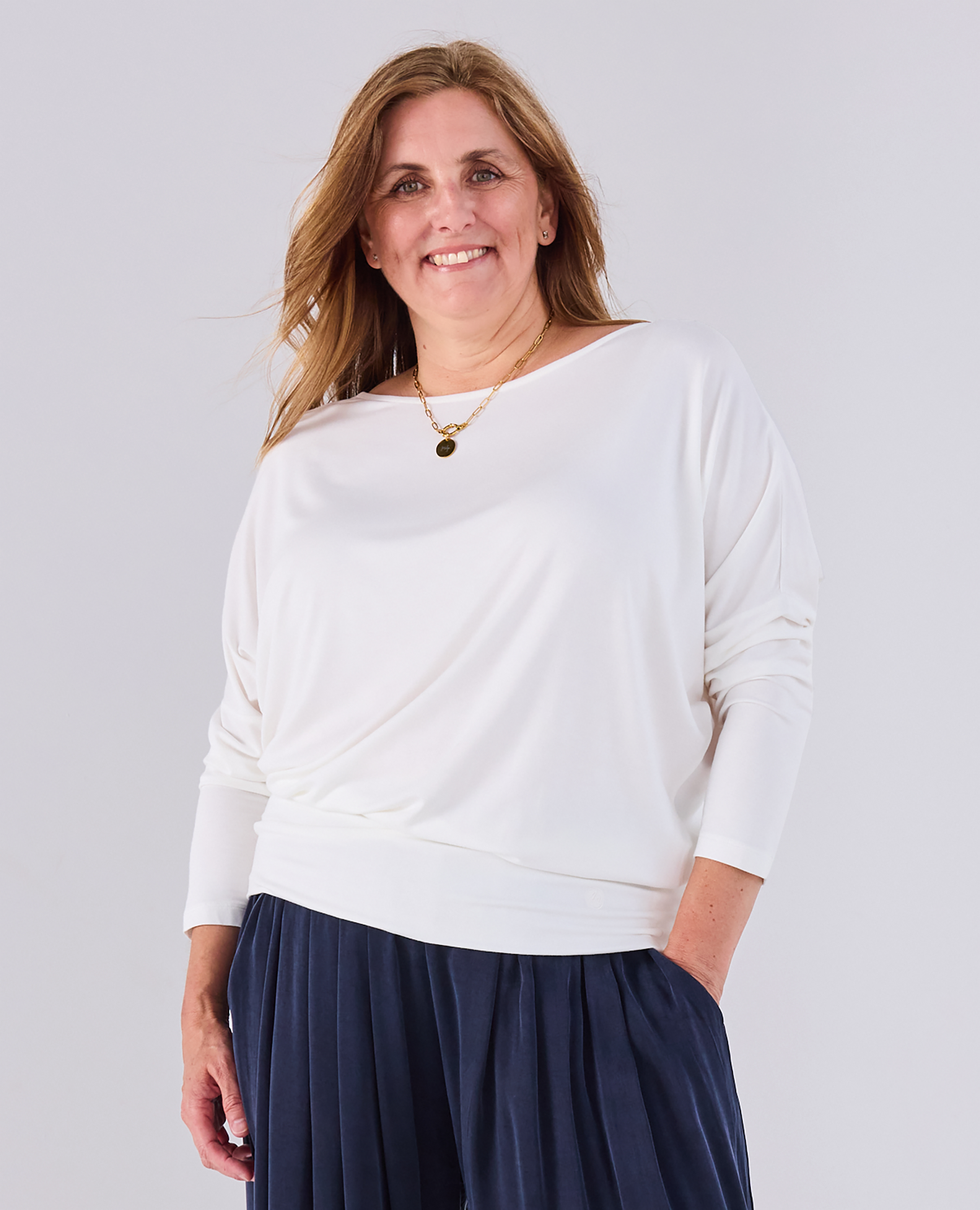A woman wearing a Melyr white Aimee long-sleeve top with a draped neckline and navy blue pleated pants, standing against a light gray studio background.