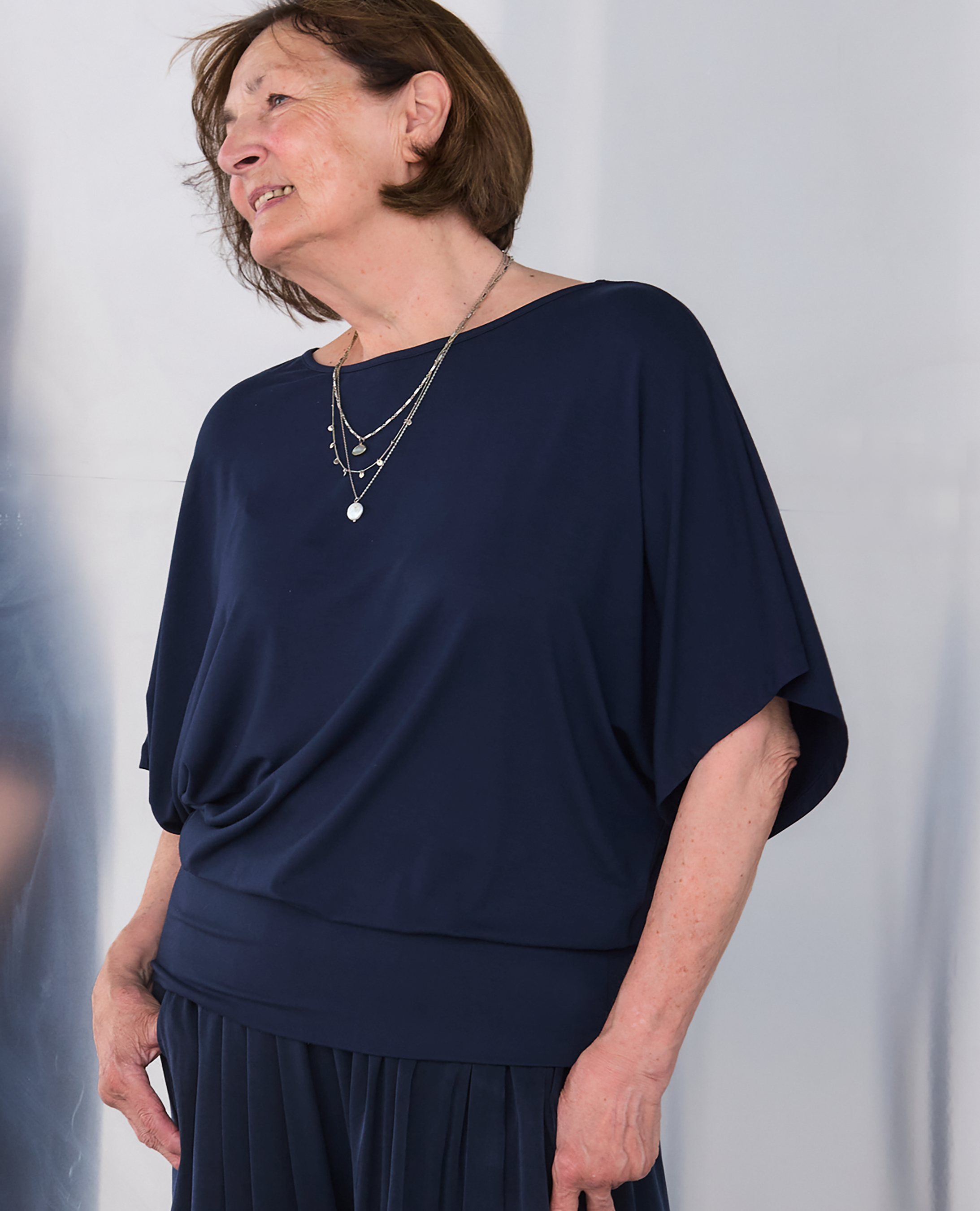 A mature woman wearing a Melyr navy Nico draped short-sleeve top and matching pleated skirt, accessorized with layered silver necklaces, photographed against a light gray background.