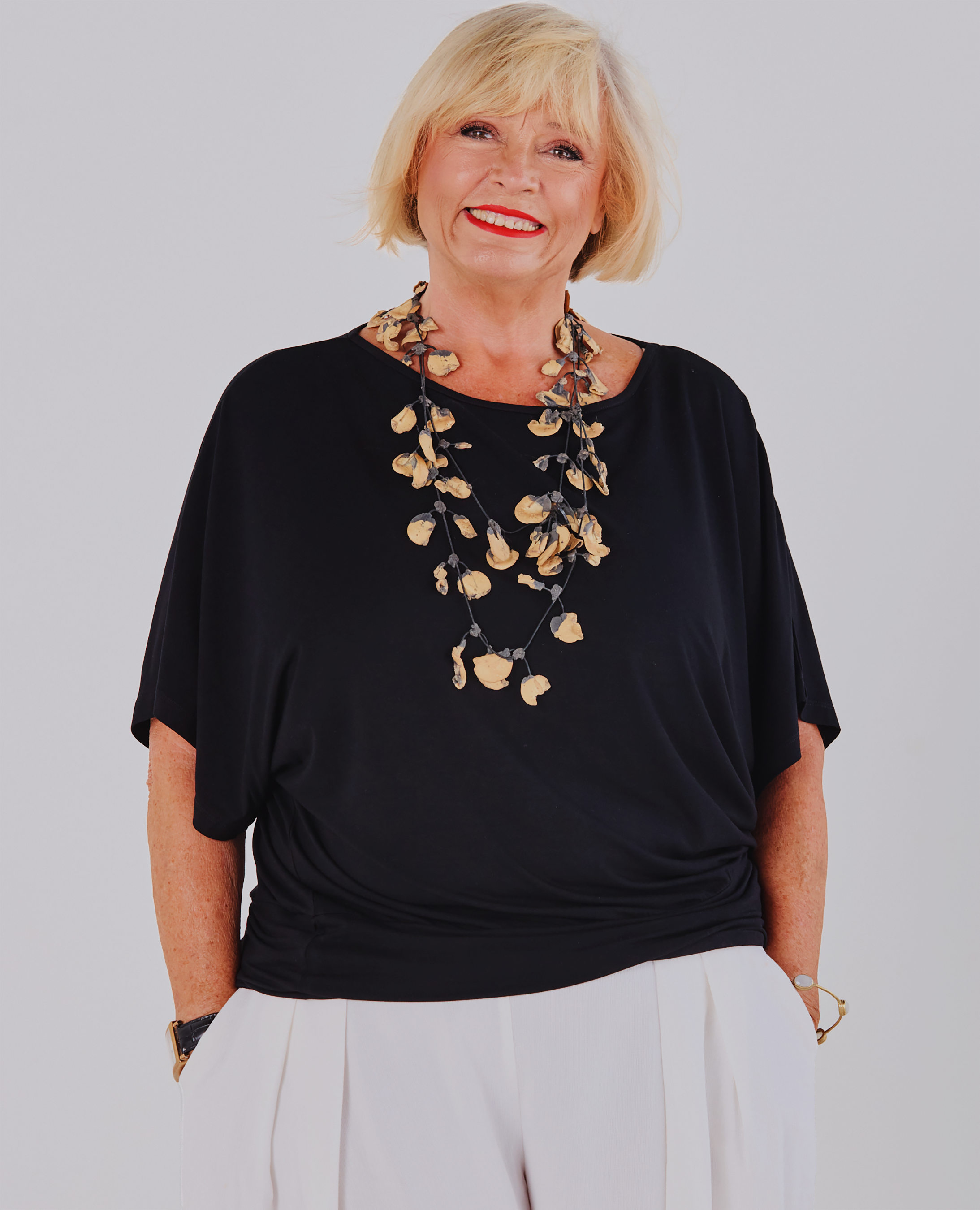 A smiling blonde woman wearing a Melyr black short-sleeve Nico top with a large layered shell necklace and white pants, photographed against a light grey studio background.