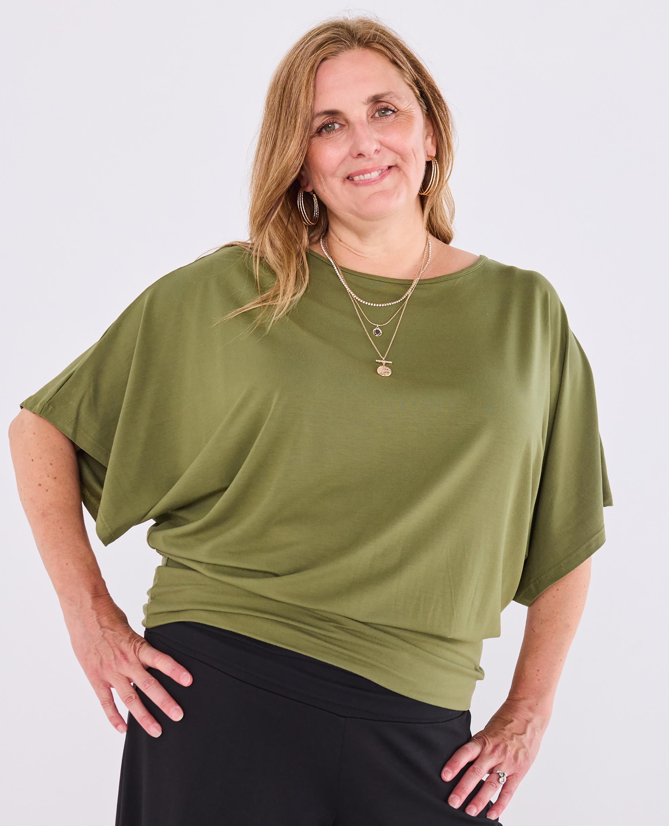 A woman modeling a Melyr, short-sleeve olive green Nico top with black bottoms, standing against a light grey studio background.