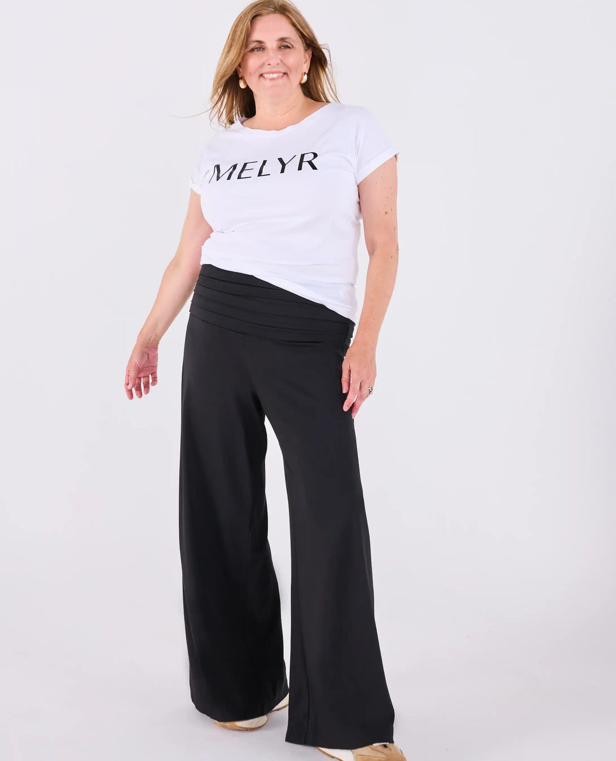 A woman modeling a white short-sleeve t-shirt with 'MELYR' printed in black across the chest, paired with black Ravello MELYRFLEX wide-leg pants, against a plain white studio background.