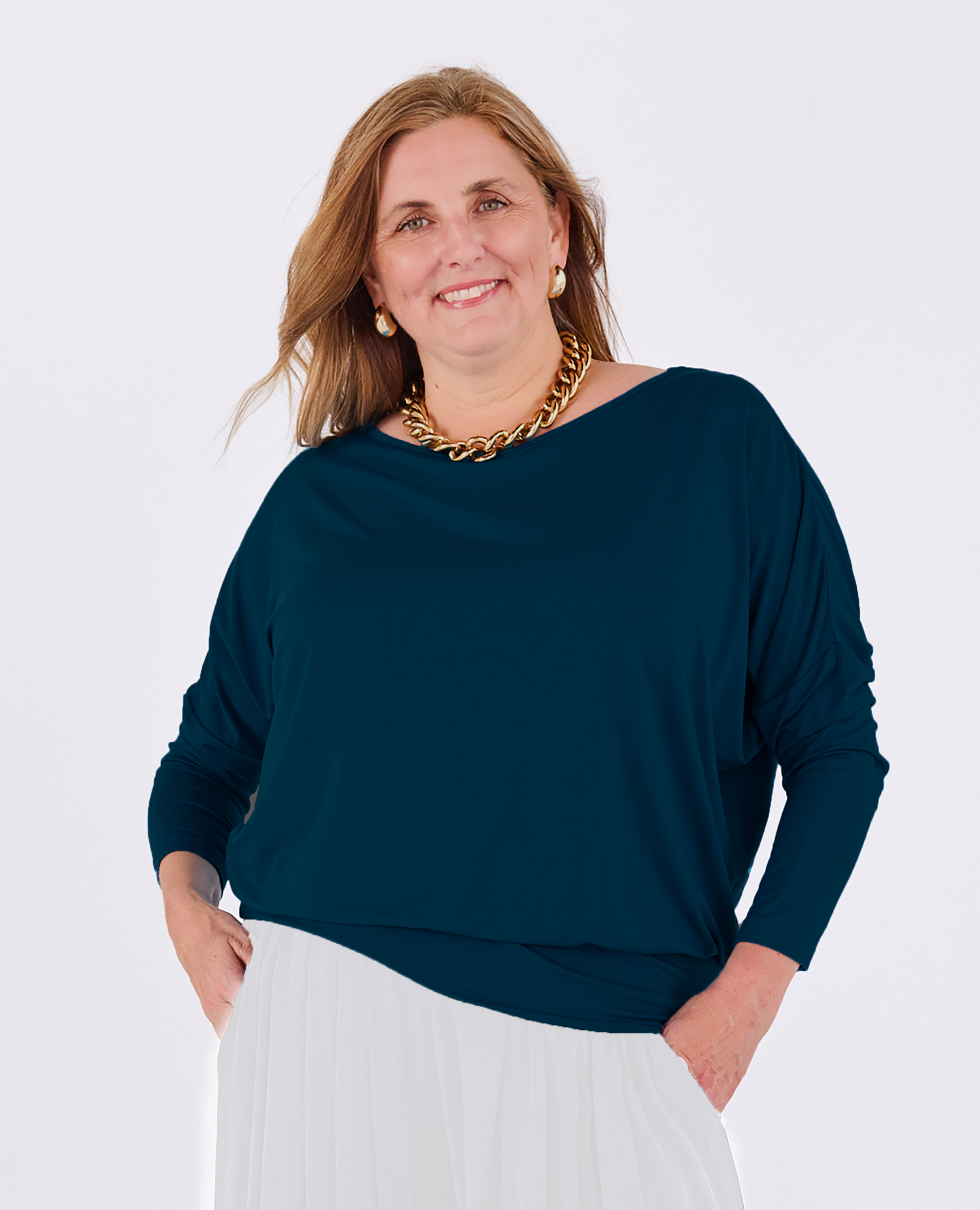 A woman modeling a dark teal long-sleeve Melyr Aimee top with a relaxed fit, paired with a white skirt, against a plain light grey background.