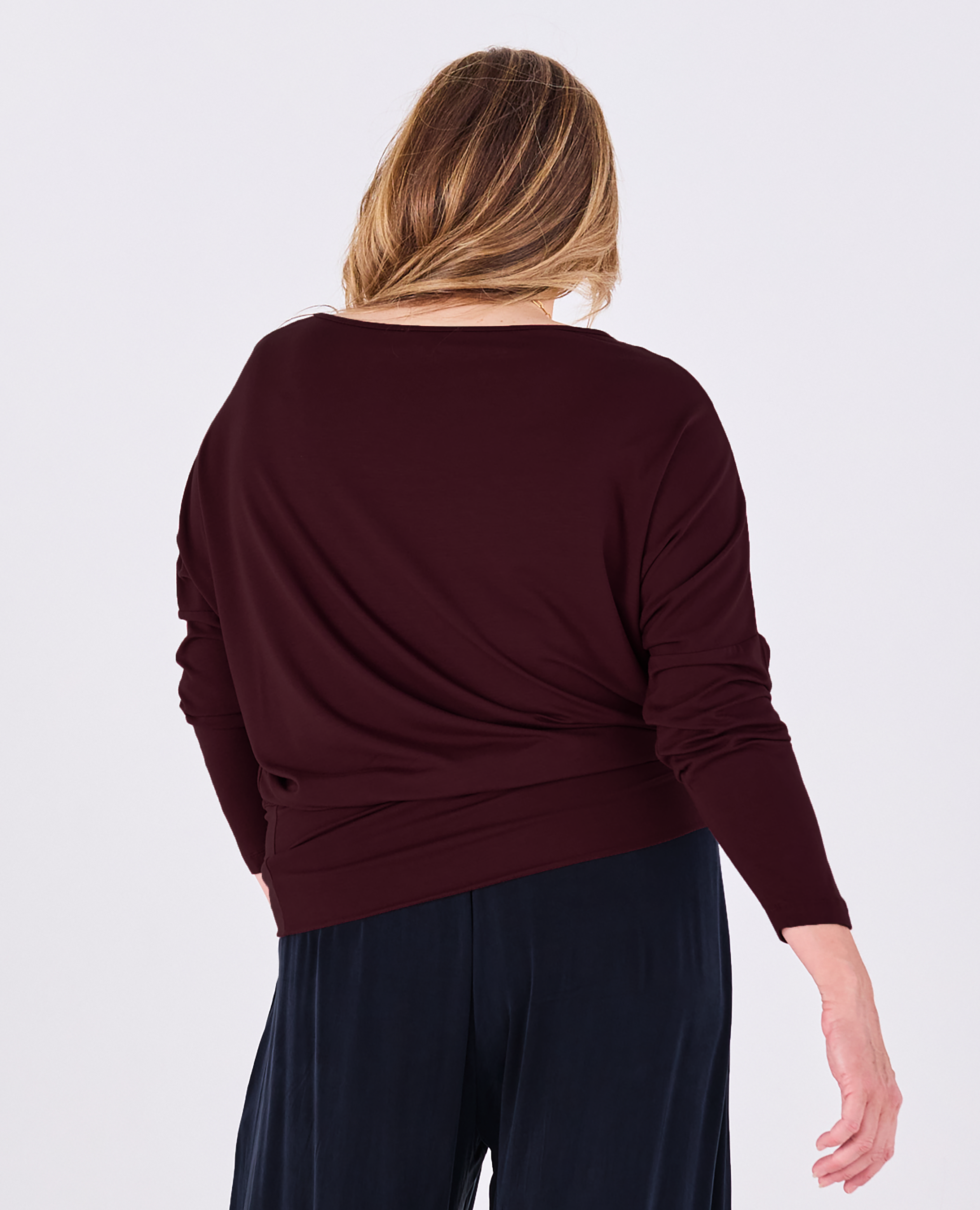 A woman viewed from the back wearing a dark burgundy long-sleeve Melyr Aimee batwing top and dark navy pants against a light grey studio background.