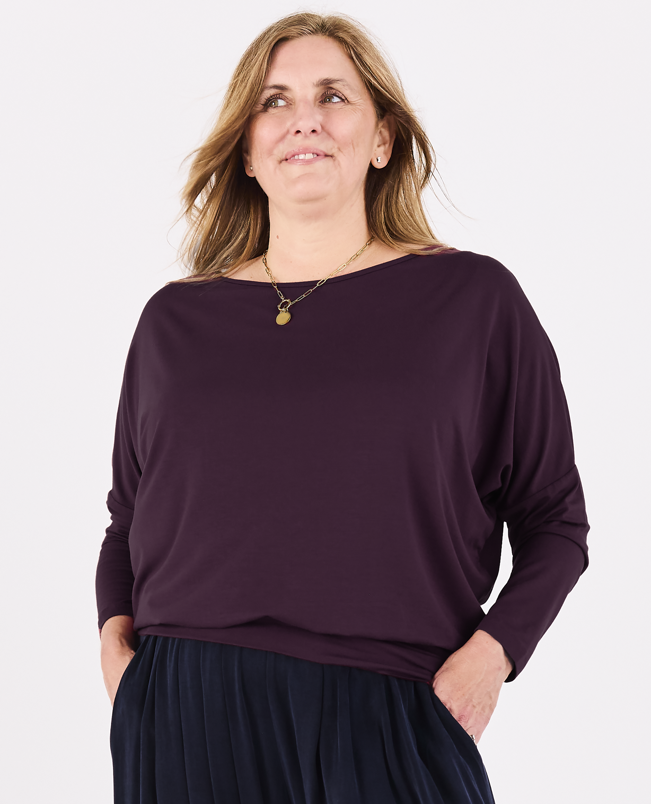 A woman wearing a dark plum Melyr Aimee long-sleeve batwing top and a navy blue pants, standing against a plain white studio background.