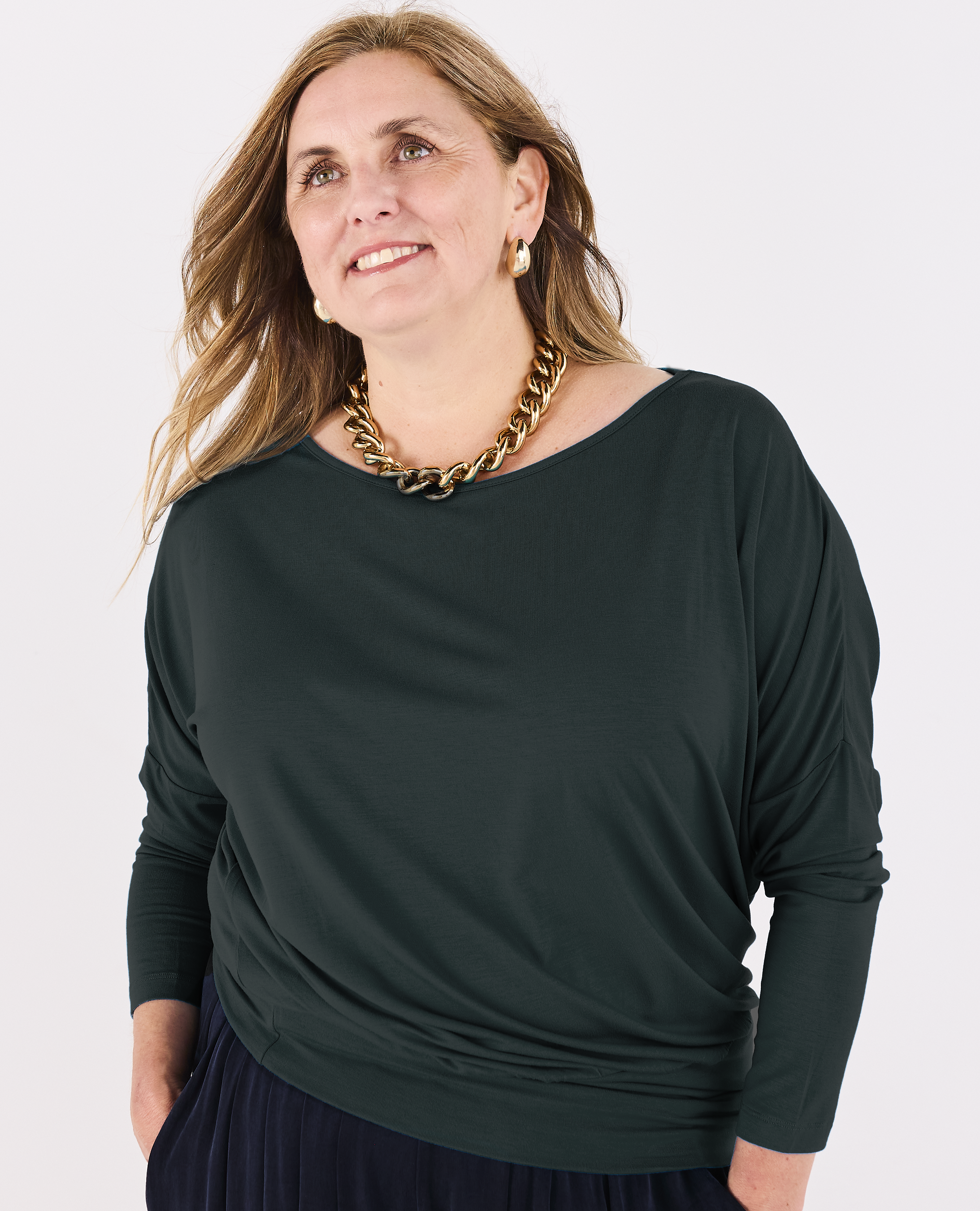 A woman wearing a dark green Melyr Aimee long-sleeve top with a wide boat neckline, styled with a chunky gold chain necklace and a dark skirt, against a plain white background.