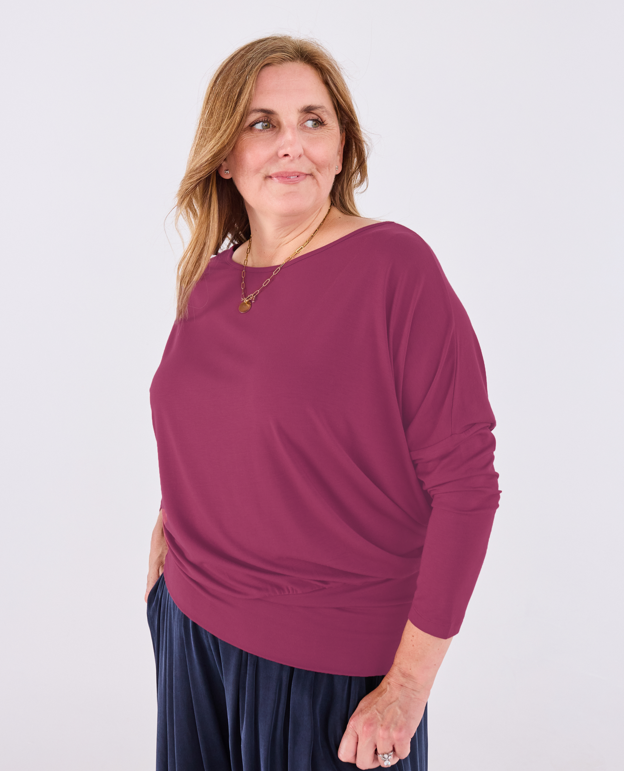A woman modeling a Melyr Aimee long-sleeve, boat-neck knit top in a deep berry purple colour, paired with dark blue pleated pants, against a plain light grey background.