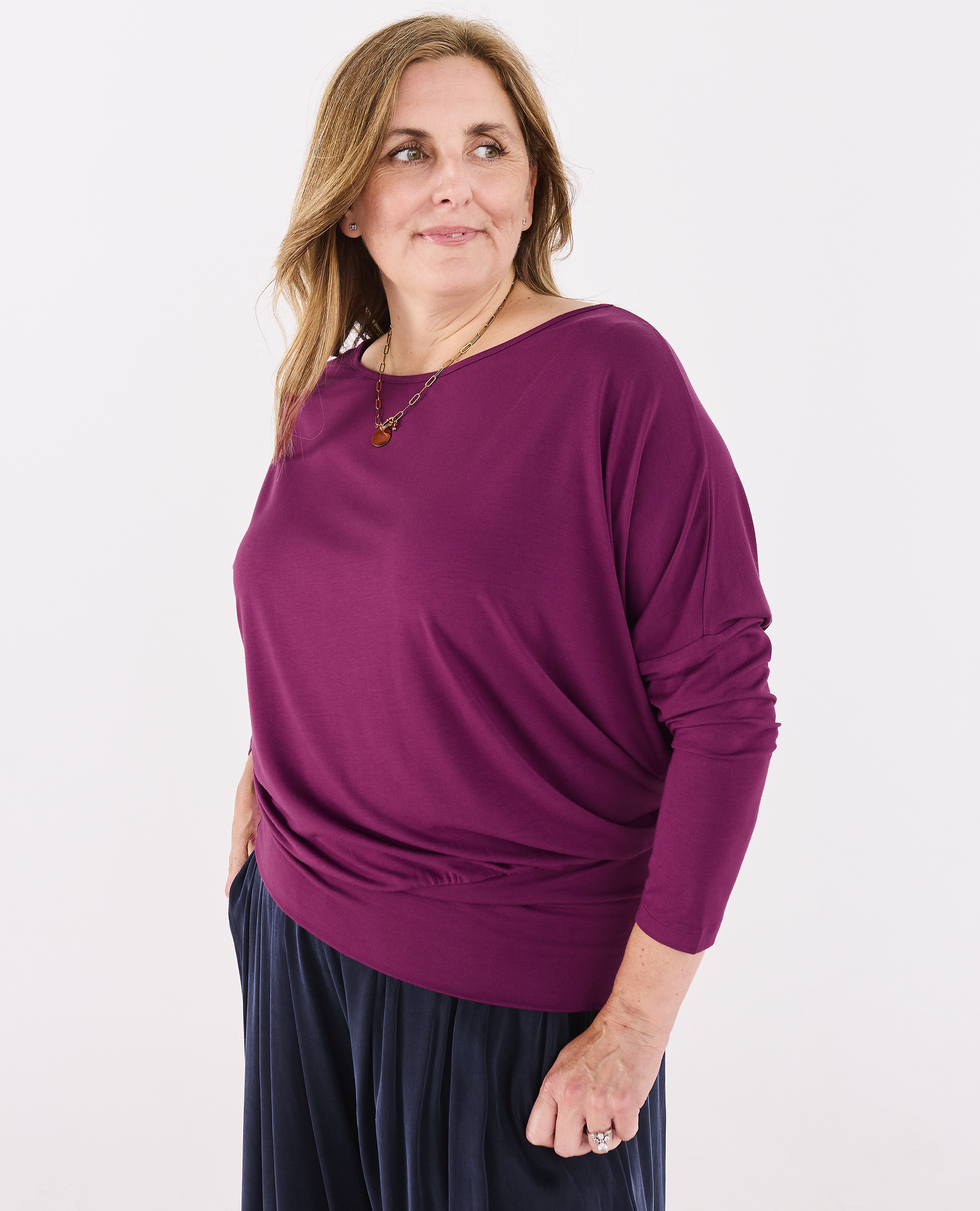 A woman modeling a Melyr Aimee long-sleeve, boat-neck knit top in a deep raspberry colour, paired with dark navy pleated pants, against a plain white studio background.