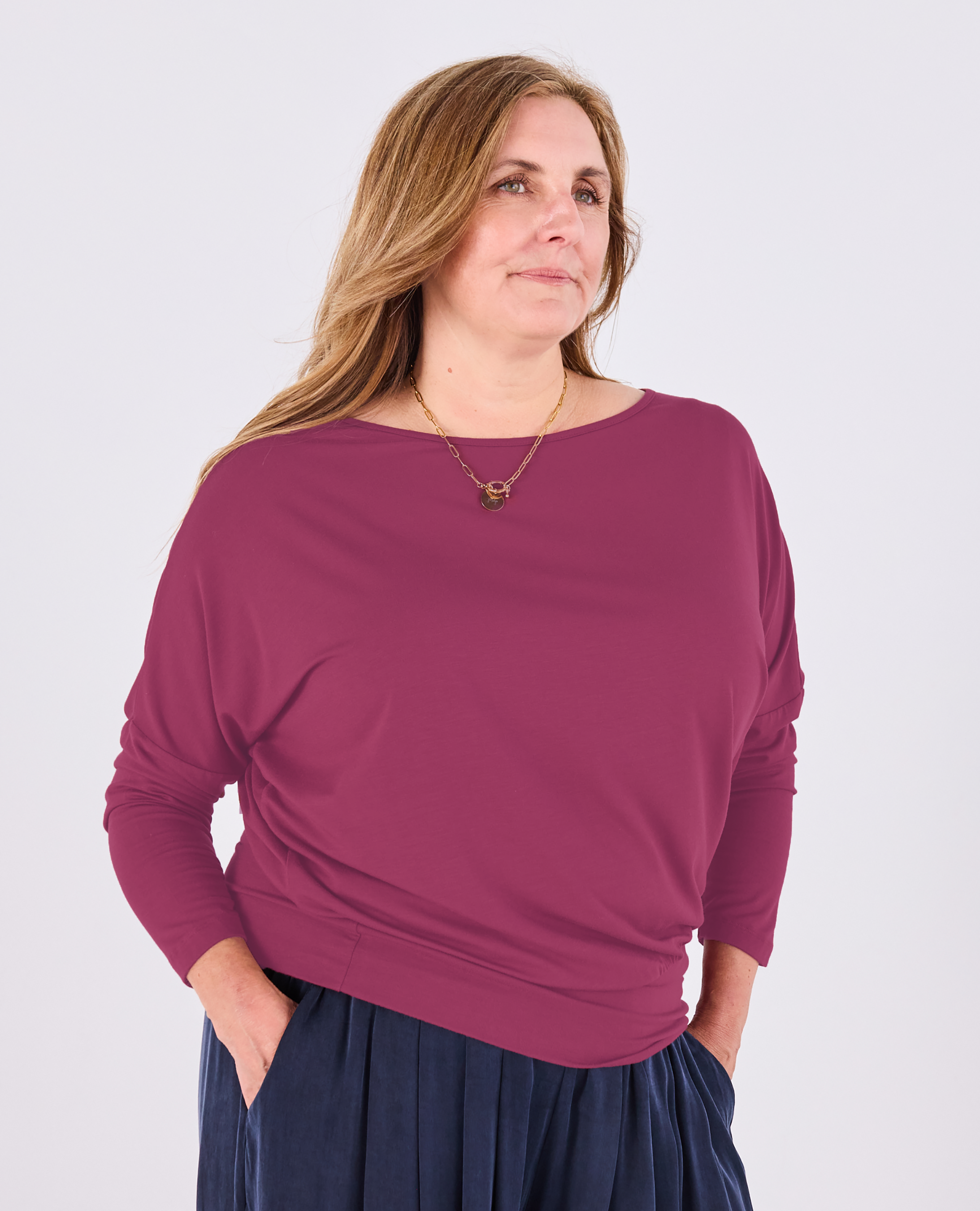 A mature woman wearing a Melyr Aimee long-sleeve batwing top in a raspberry colour, paired with navy blue pants, against a plain light grey background.