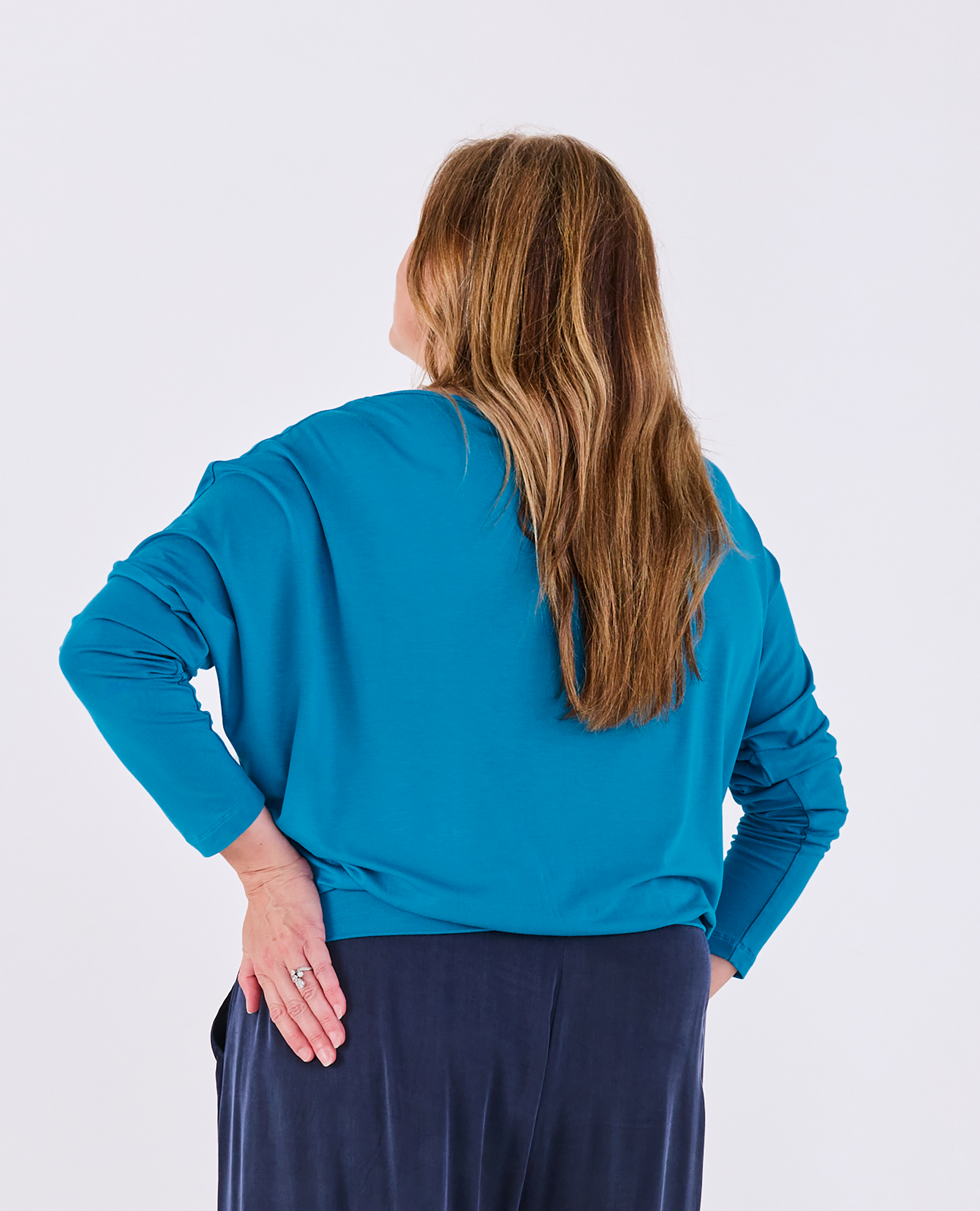 A woman with long light brown hair is shown from behind wearing a bright teal blue long-sleeve Melyr Aimee batwing top and navy blue pants against a plain light grey studio background.