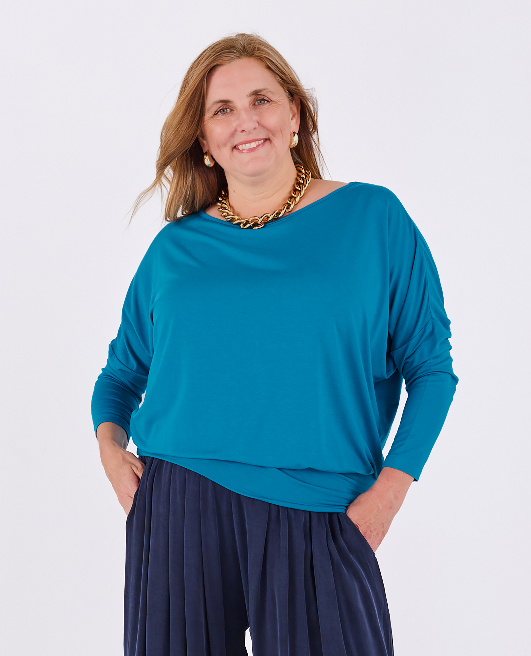 A woman modeling a Melyr teal blue long-sleeve boat-neck Aimee top with dark navy pleated pants, accessorized with a chunky gold chain necklace, against a plain white studio background.