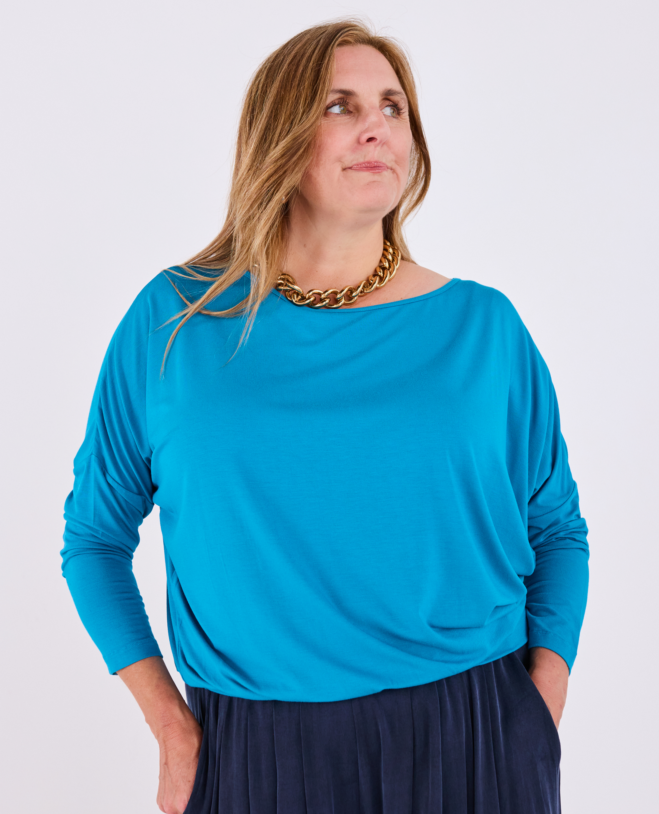 A woman wearing a Melyr teal blue long-sleeve Aimee top with a wide boat neckline, paired with a dark navy skirt and a chunky gold chain necklace, photographed against a light gray studio background.