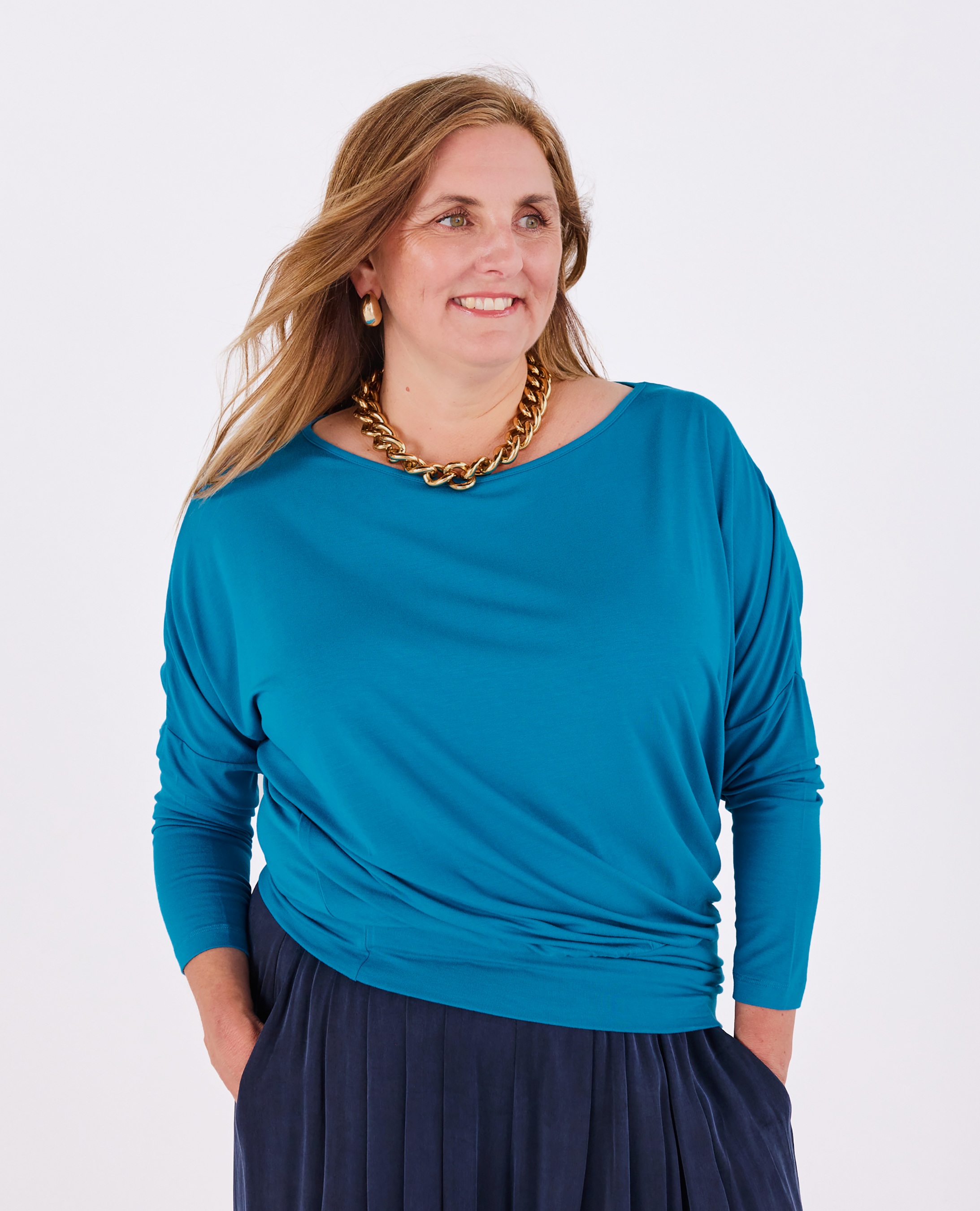 A woman wearing a Melyr teal long-sleeve Aimee top and a navy pleated skirt, accessorized with a chunky gold chain necklace, standing against a light gray studio background.