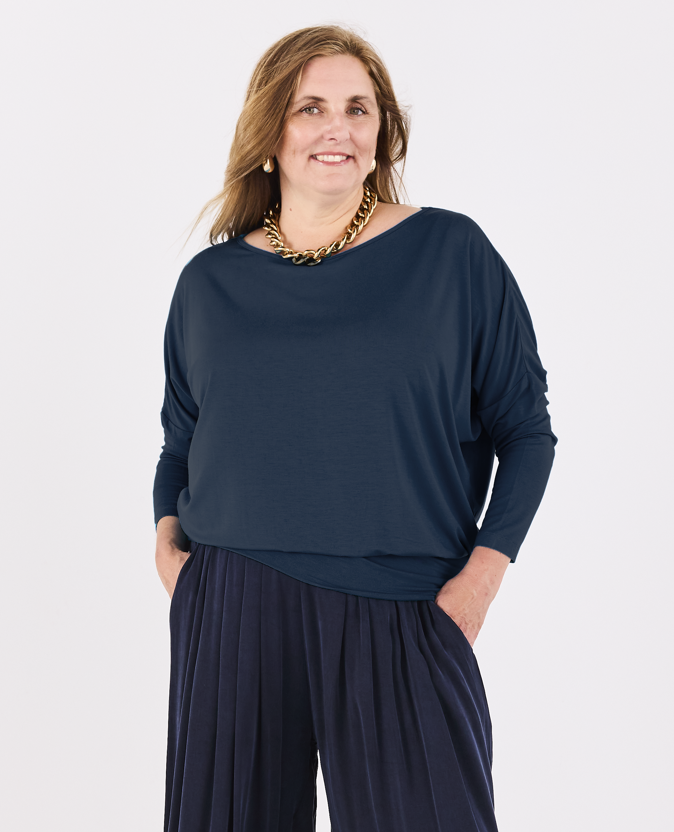 A woman modeling a Melyr Aimee teal blue long-sleeve batwing top with matching pleated navy pants, accessorised with a chunky gold chain necklace, against a plain white studio background.