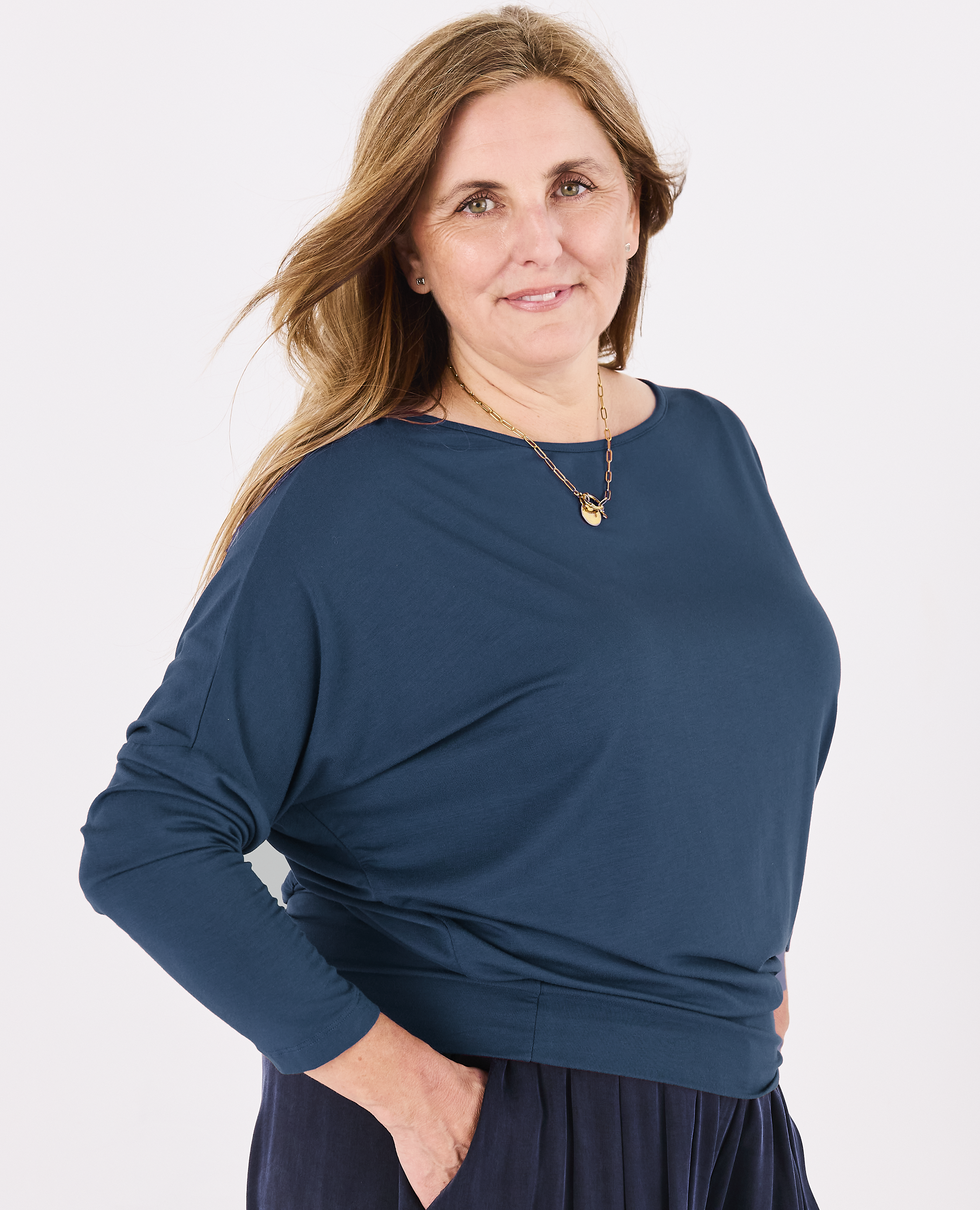 A woman wearing a Melyr dark teal blue long-sleeve Aimee batwing top and a navy skirt, standing against a plain white studio background.