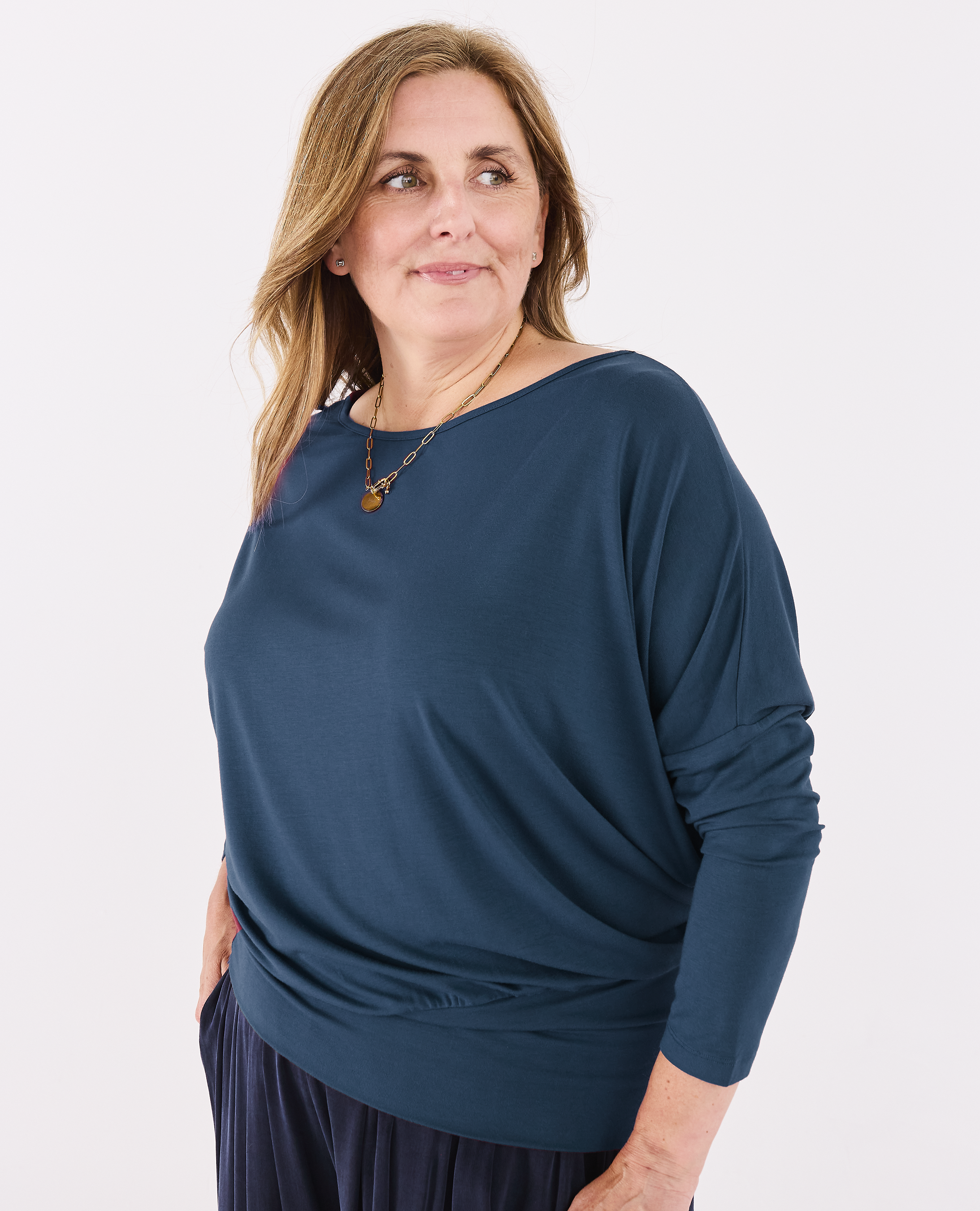 A woman wearing a Melyr dark teal blue batwing top with a wide boat neckline, paired with navy blue pleated pants, photographed against a plain white studio background.