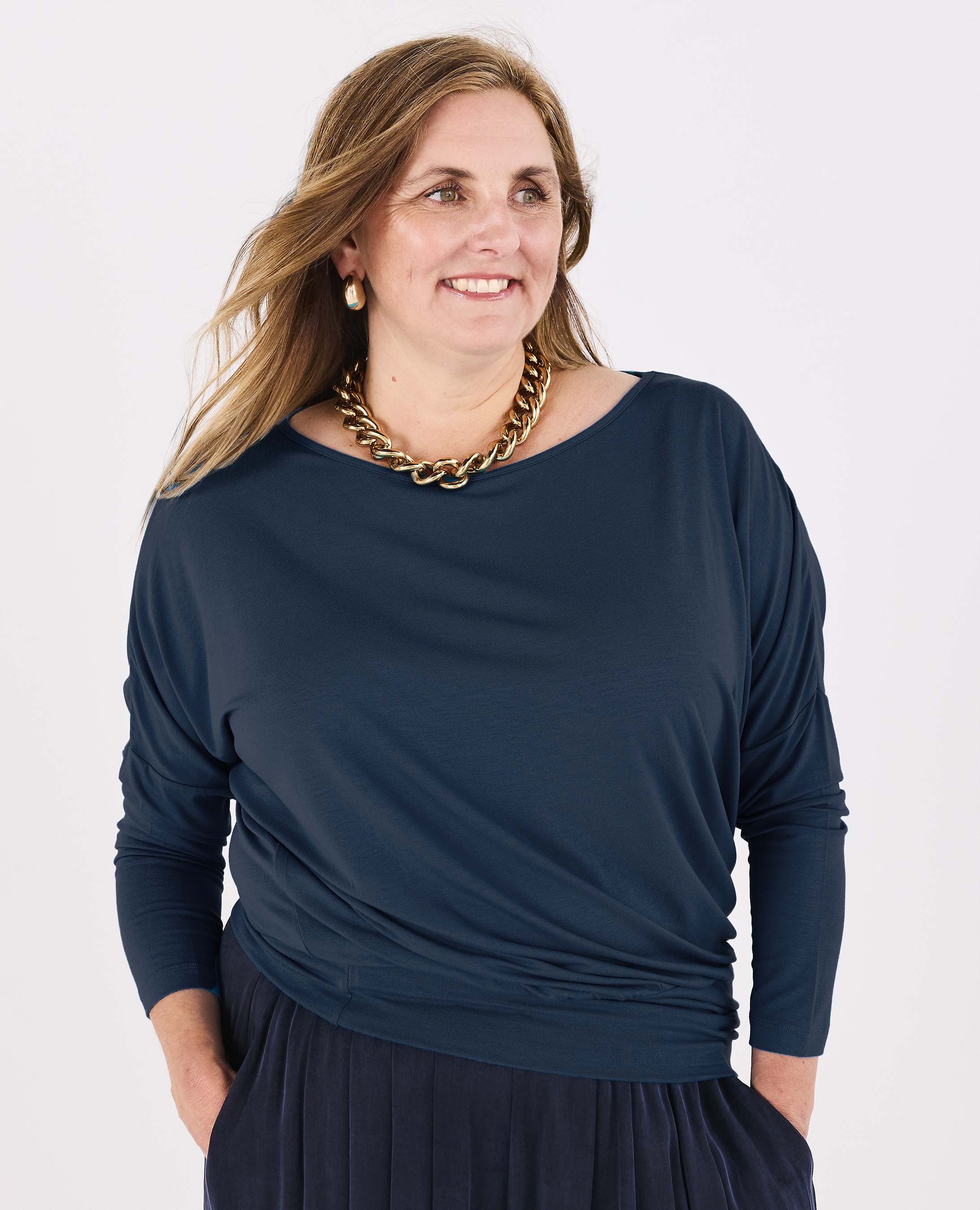 A woman wearing a Melyr teal blue long-sleeve batwing top with a draped, boat-style neckline and a gathered waist, paired with a pleated navy pant and a chunky gold chain necklace, against a plain white background.
