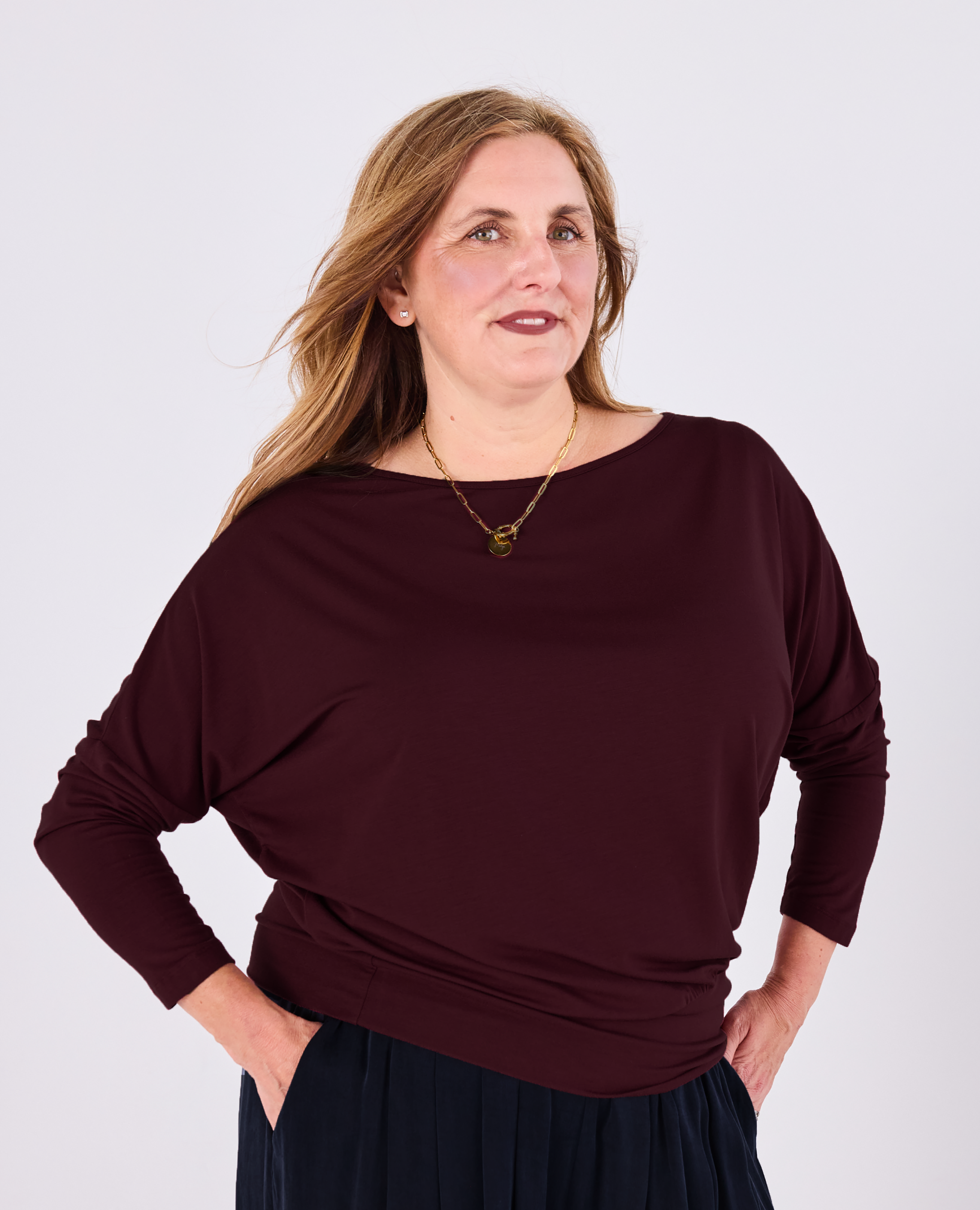 A woman wearing a dark burgundy long-sleeve Melyr Aimee batwing top and a dark navy skirt, standing against a light grey studio background.
