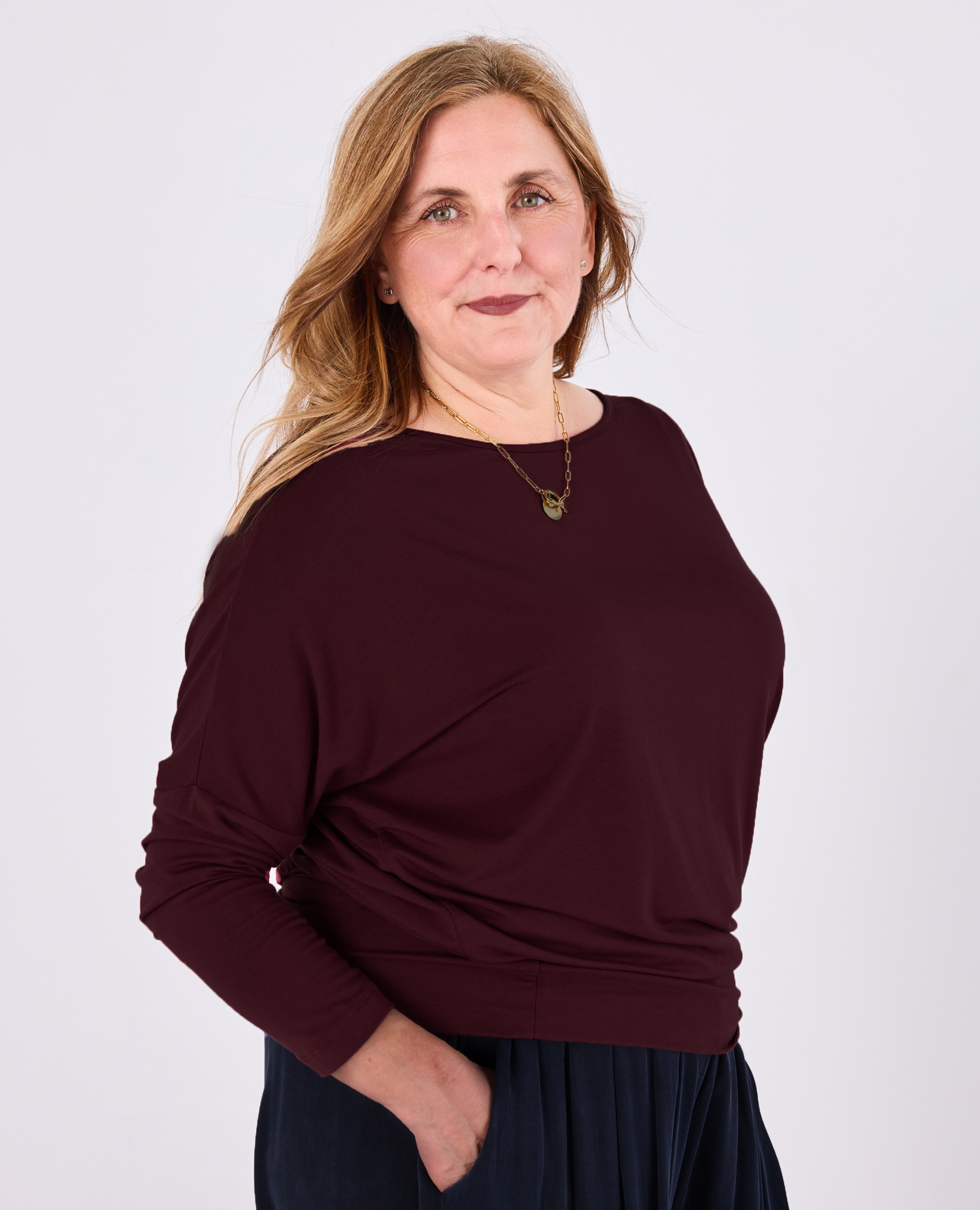 A woman modeling a long-sleeve boat-neck batwing top by Melyr in deep burgundy, paired with a dark navy skirt, against a plain light grey studio background.