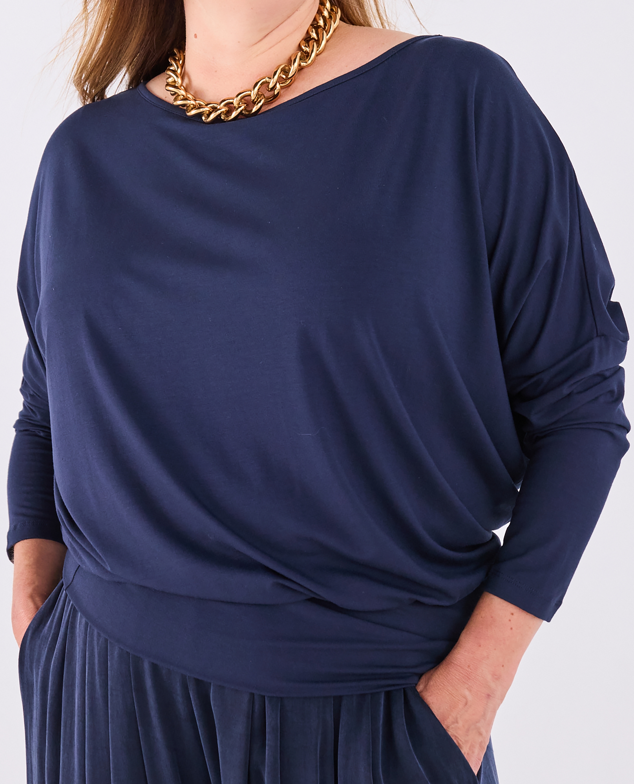 A close-up of a woman wearing a Melyr navy Aimee draped long-sleeve batwing top with a boat neckline, styled with a chunky gold chain necklace.