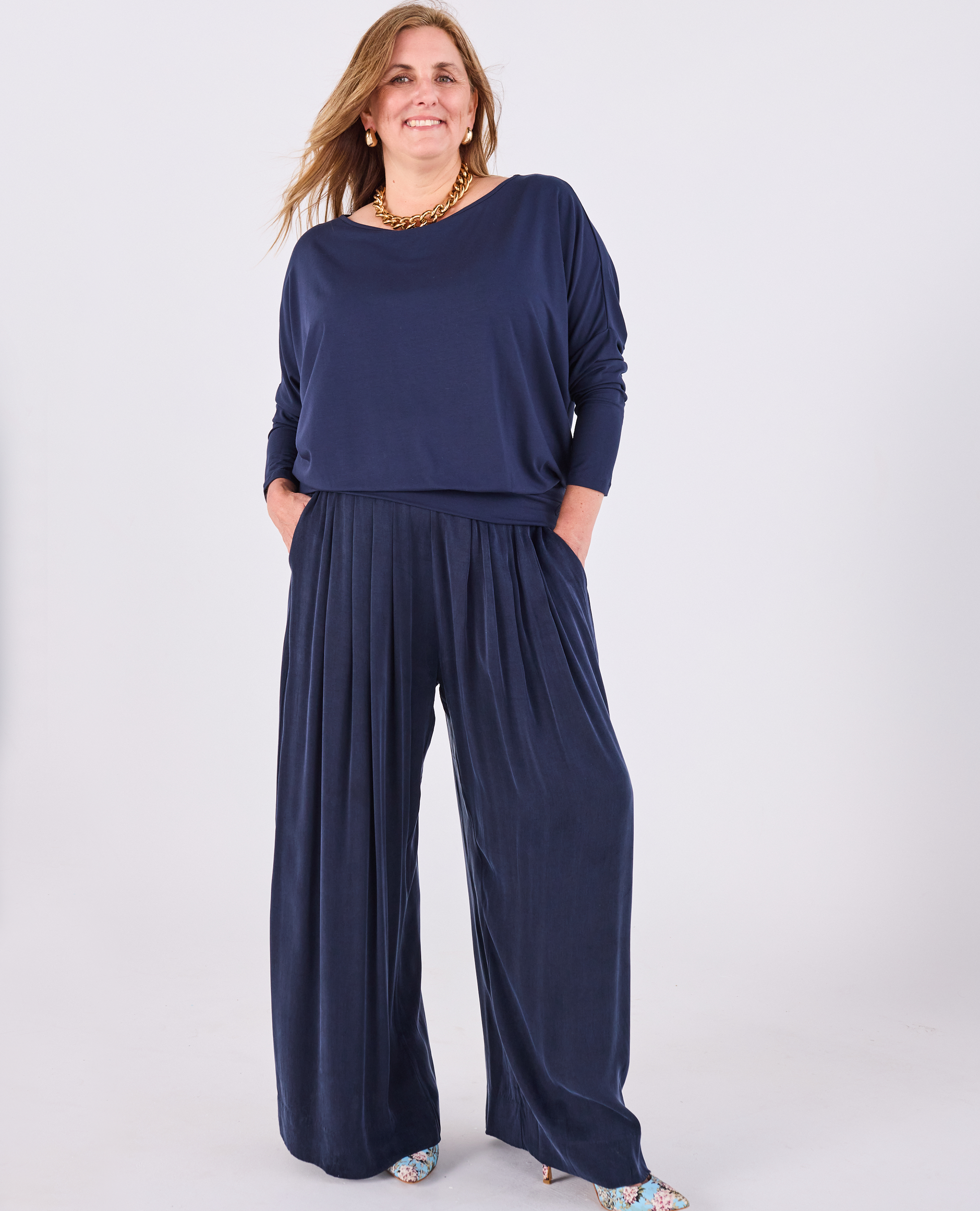 A woman modeling a Melyr navy Aimee long-sleeve top and Melyr Portofino wide-leg pleated pants set against a plain light grey studio background.