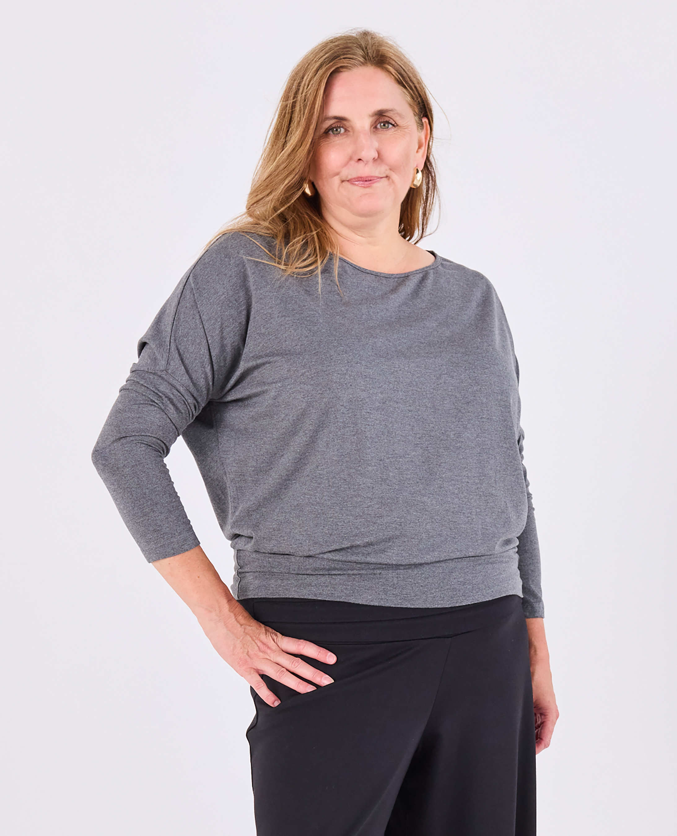 A woman modeling a heather gray long-sleeve Melyr top with a boat neckline and black pants against a light gray studio background.