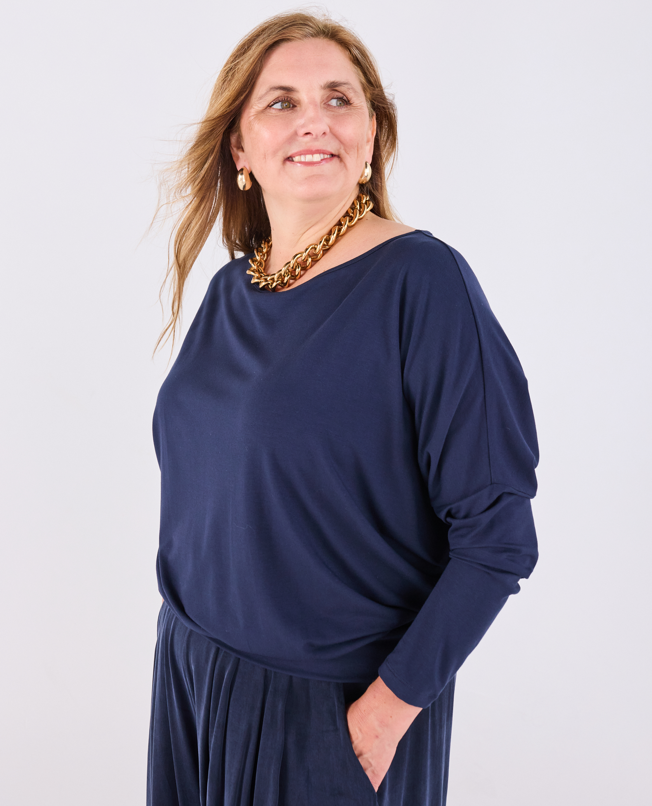 A woman wearing a Melyr navy Aimee long-sleeve batwing top and Portofino pleated trousers, accessorized with a chunky gold chain necklace, photographed against a light grey studio background
