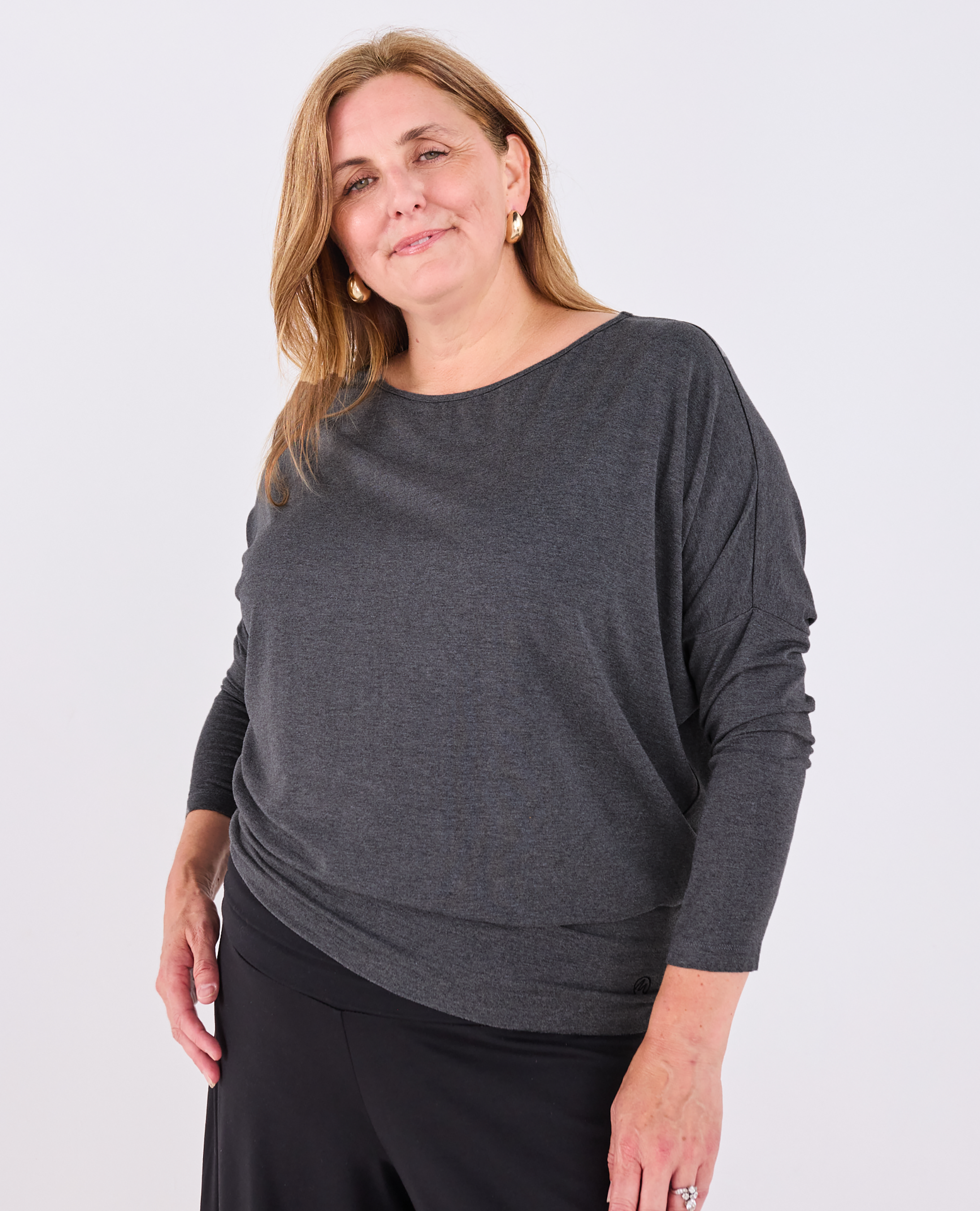 A woman modeling a dark gray heathered long-sleeve Melyr Aimee top with a wide boat neckline, paired with black pants, against a light gray studio background.