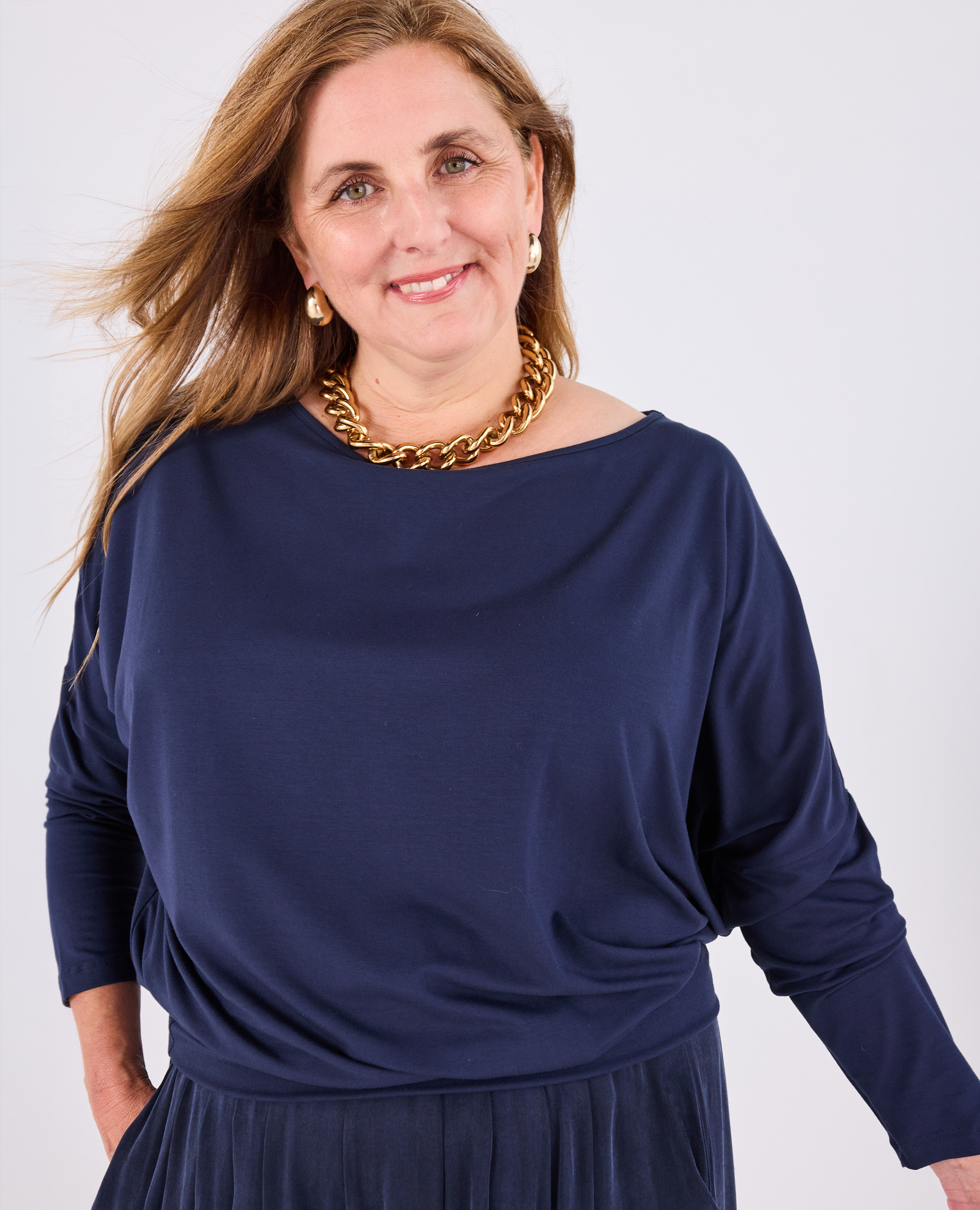 A woman wearing a Melyr navy Aimee long-sleeve top with a boat neckline and a chunky gold chain necklace, photographed against a light grey studio background.