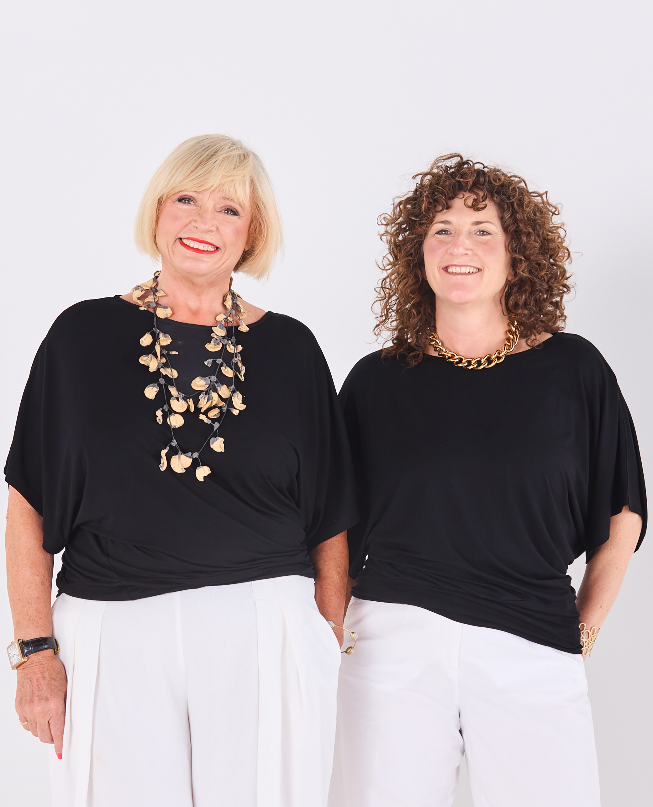 Two mature women standing side by side against a plain light grey studio background. Both are wearing Melyr Nico black short-sleeve tops and white pants, with one wearing a long layered shell necklace and the other a chunky gold chain necklace.