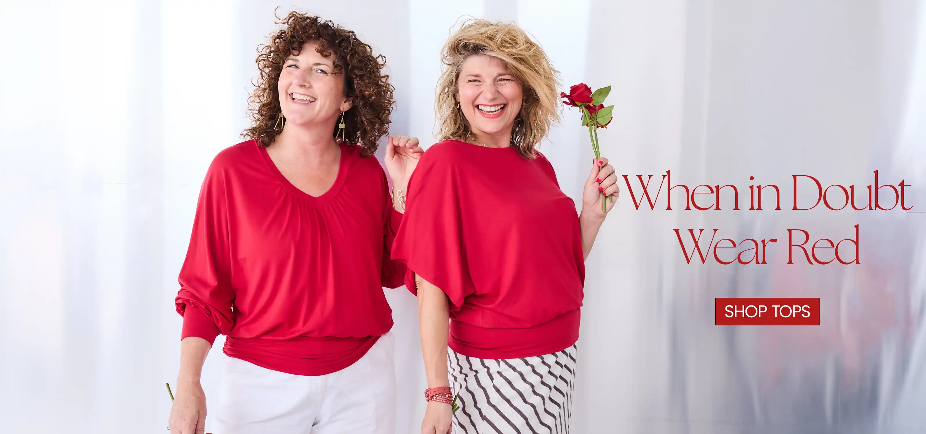 Two smiling women wearing matching bright red Melyr tops pose together against a white curtain backdrop. One holds a single red rose, and the right side includes the text 'When in Doubt Wear Red' with a 'SHOP TOPS' button.