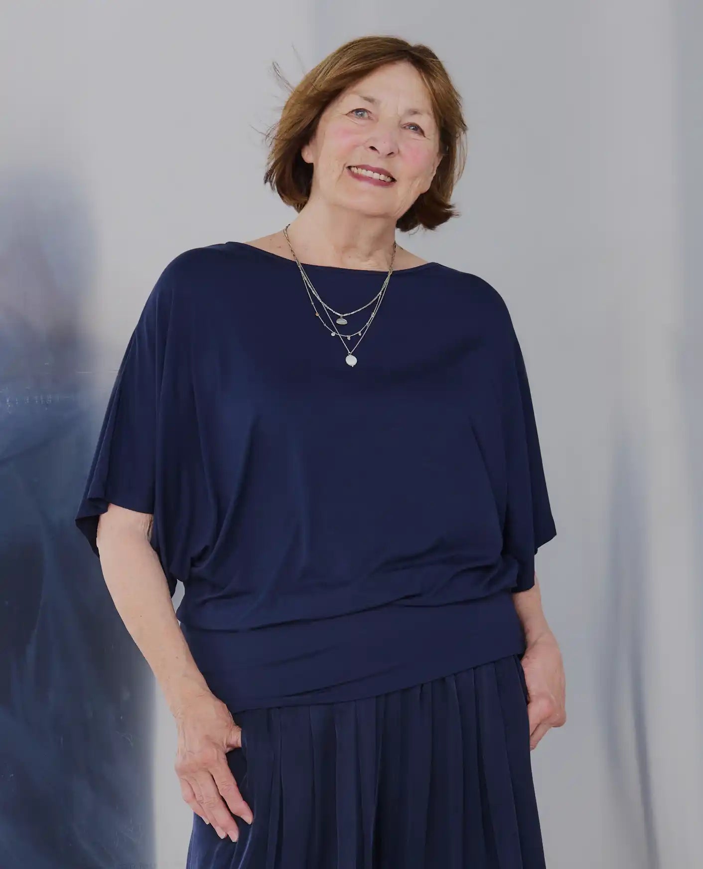 A mature woman with short brown hair wearing a Melyr navy Nico draped short-sleeve top and matching pleated skirt, accessorized with layered silver necklaces, standing against a light gray studio backdrop.