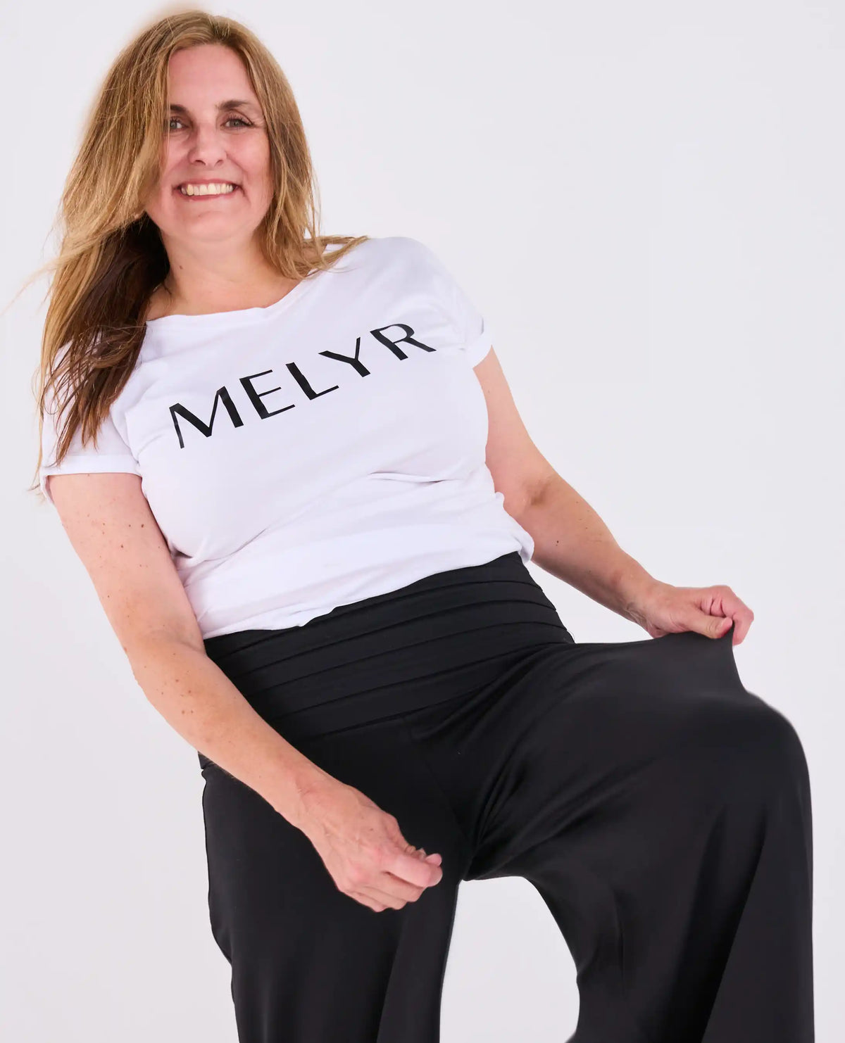 A woman wearing a white short-sleeve t-shirt with the brand name 'MELYR' printed in black across the chest, paired with black Ravello MELYRFLEX wide-leg trousers, posing against a light gray studio background.