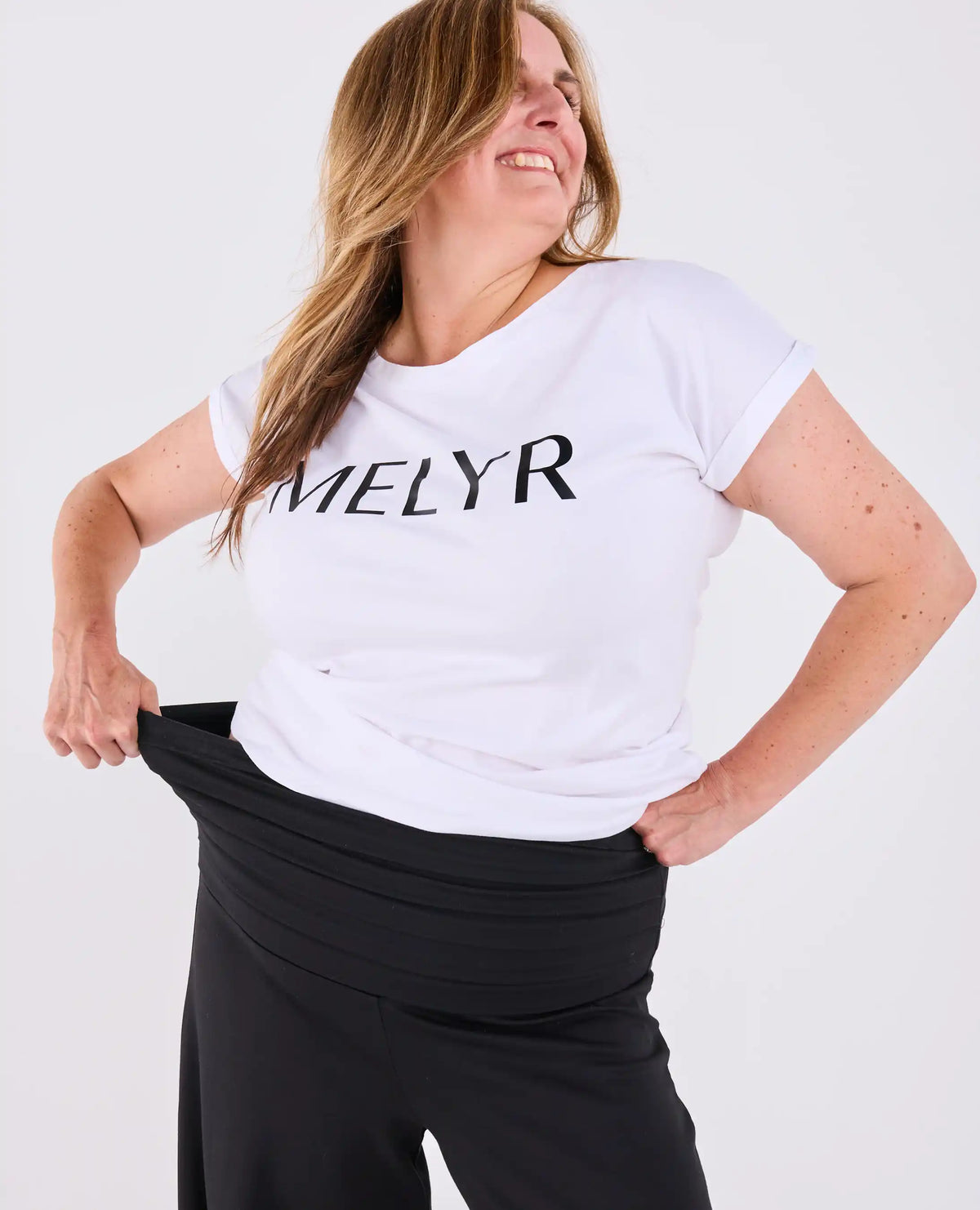 A woman wearing a white short-sleeve t-shirt with the brand name 'MELYR' printed in black across the chest, paired with black Ravello MELYRFLEX pants, posing against a light gray studio background.