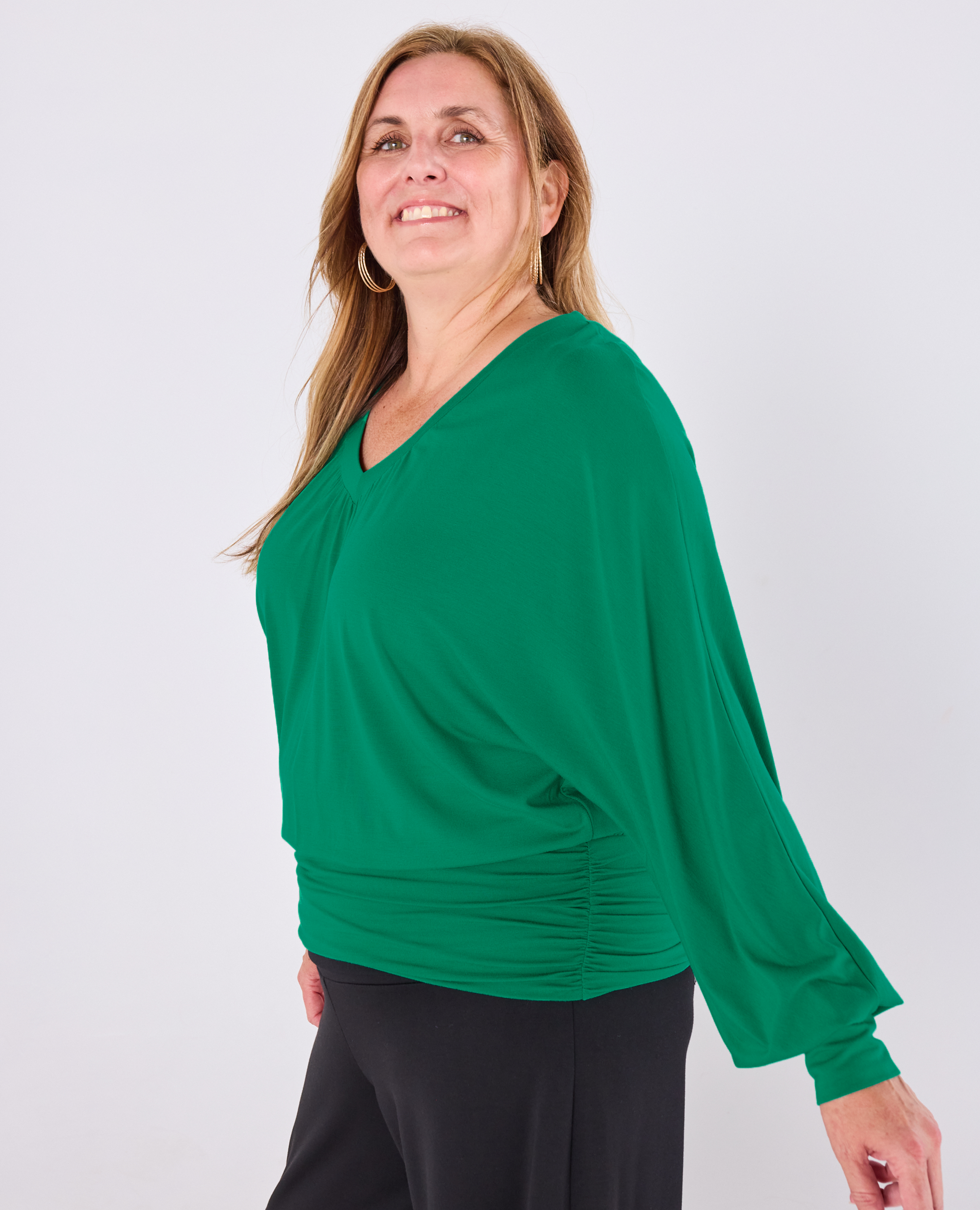 A woman wearing a vibrant green Melyr vivienne long-sleeve top with a V-neck, draped front, and gathered ruching at the waist, paired with black pants, photographed against a plain light gray background.