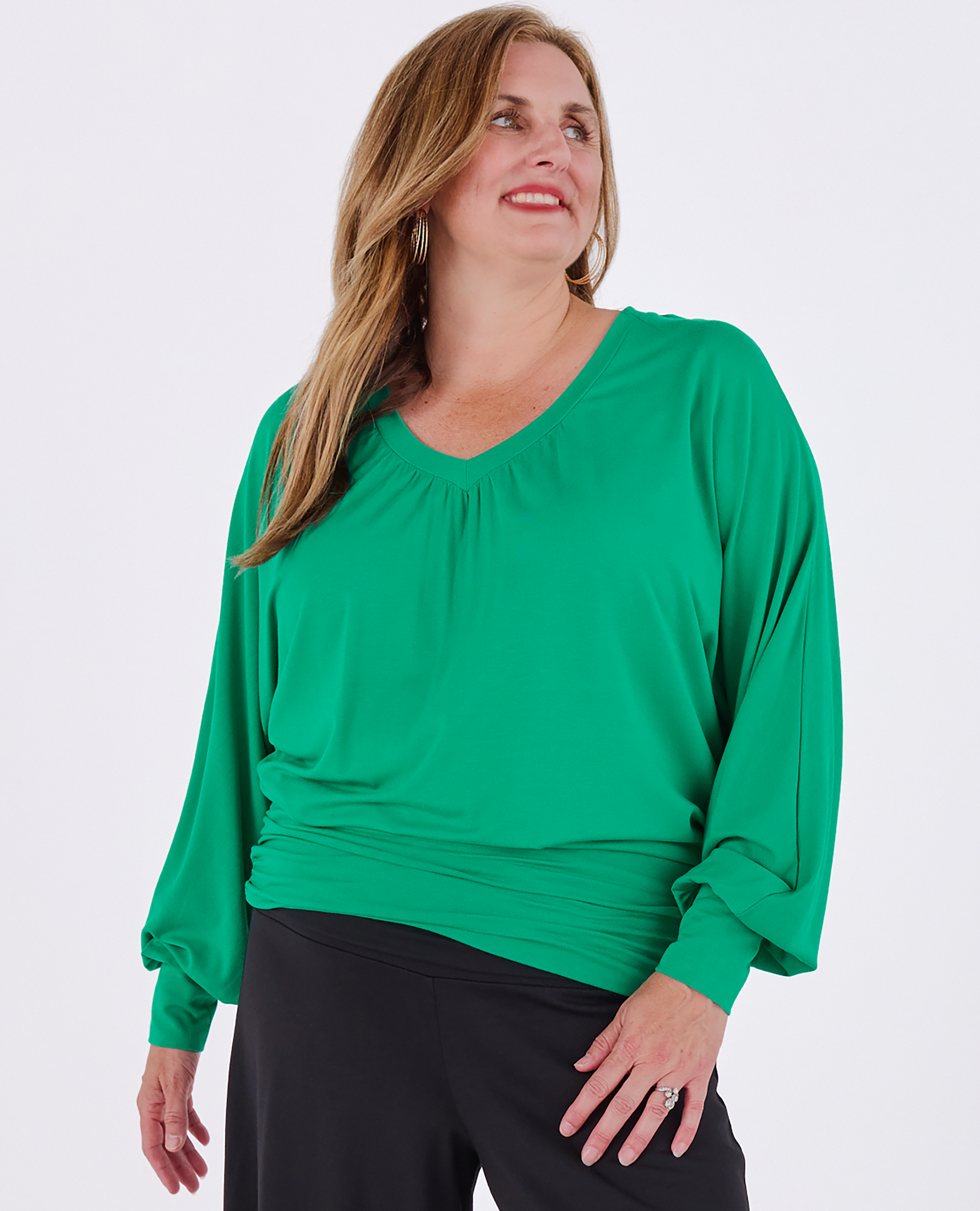 A woman wearing a emerald green Melyr vivienne long-sleeve batwing top with a gathered V-neckline and black pants, photographed against a light grey studio background.