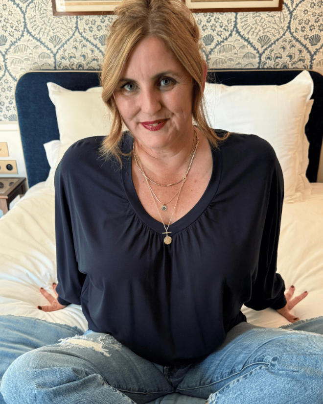 A woman with blonde hair sits cross-legged on a bed wearing a Melyr dark navy blue long-sleeve vivienne top and light blue jeans. The background shows a patterned wallpaper, a dark blue headboard, and white bedding.
