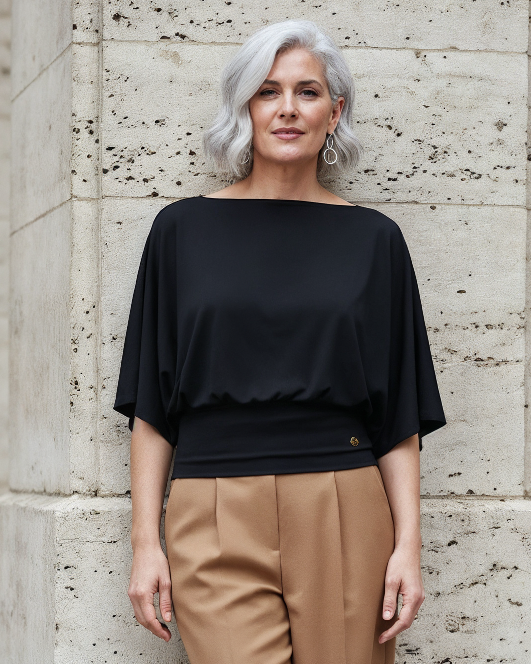 Woman wearing a black Melyr top and beige pants standing against a textured wall.