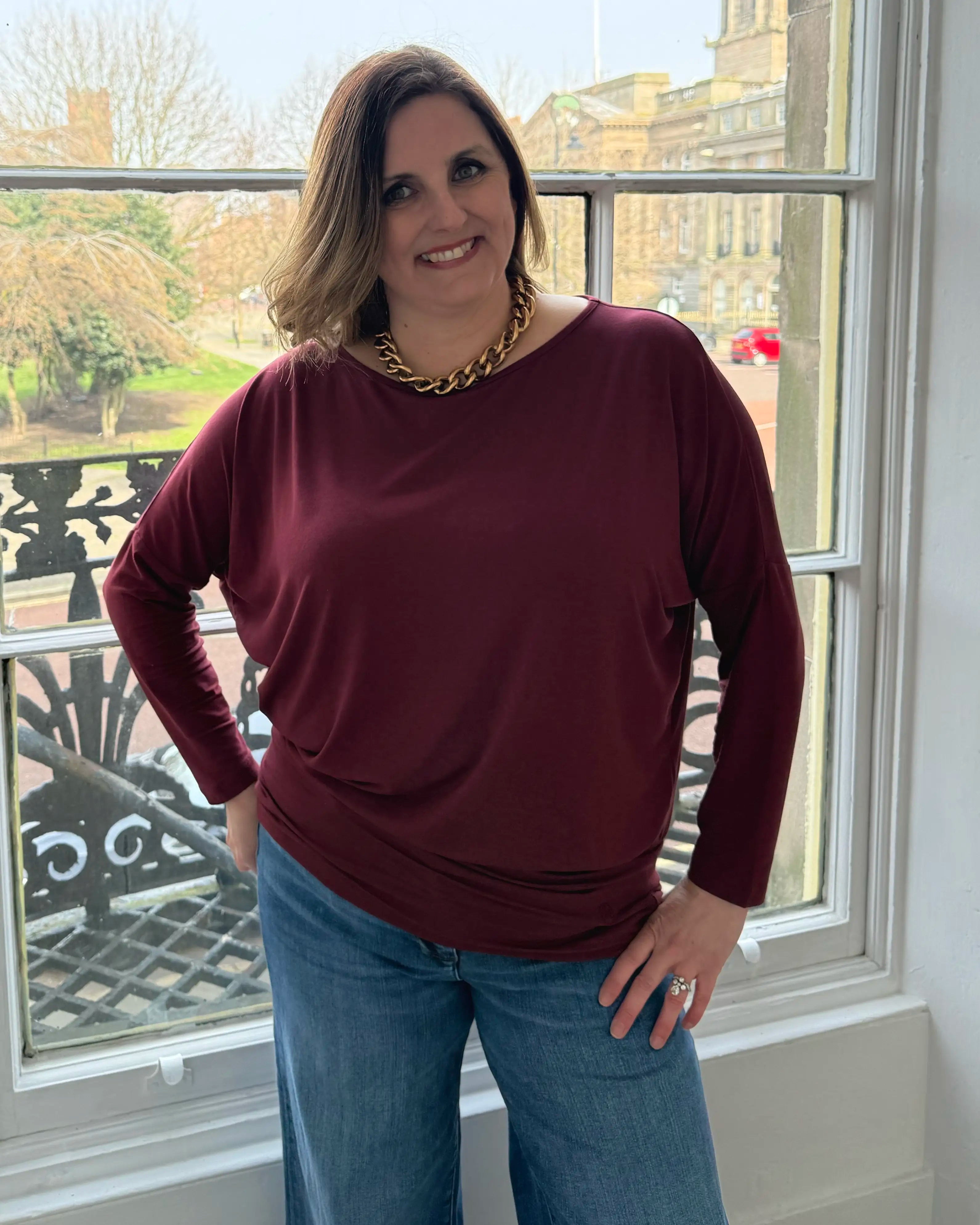 Woman wearing a Melyr burgundy long-sleeve top and blue jeans standing in front of a window with a garden view.