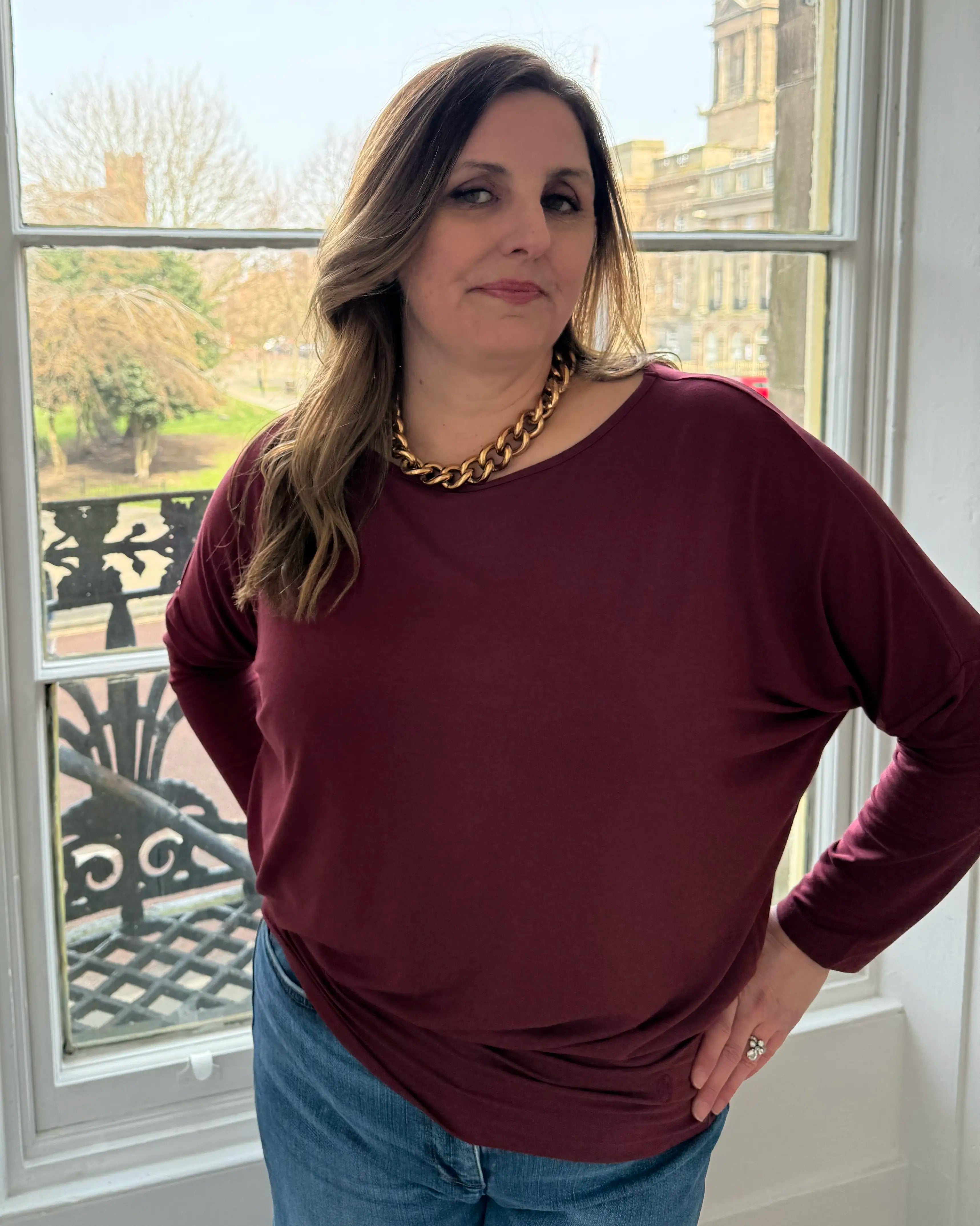 Woman wearing a burgundy sweater and blue jeans standing in front of a window with a garden view.