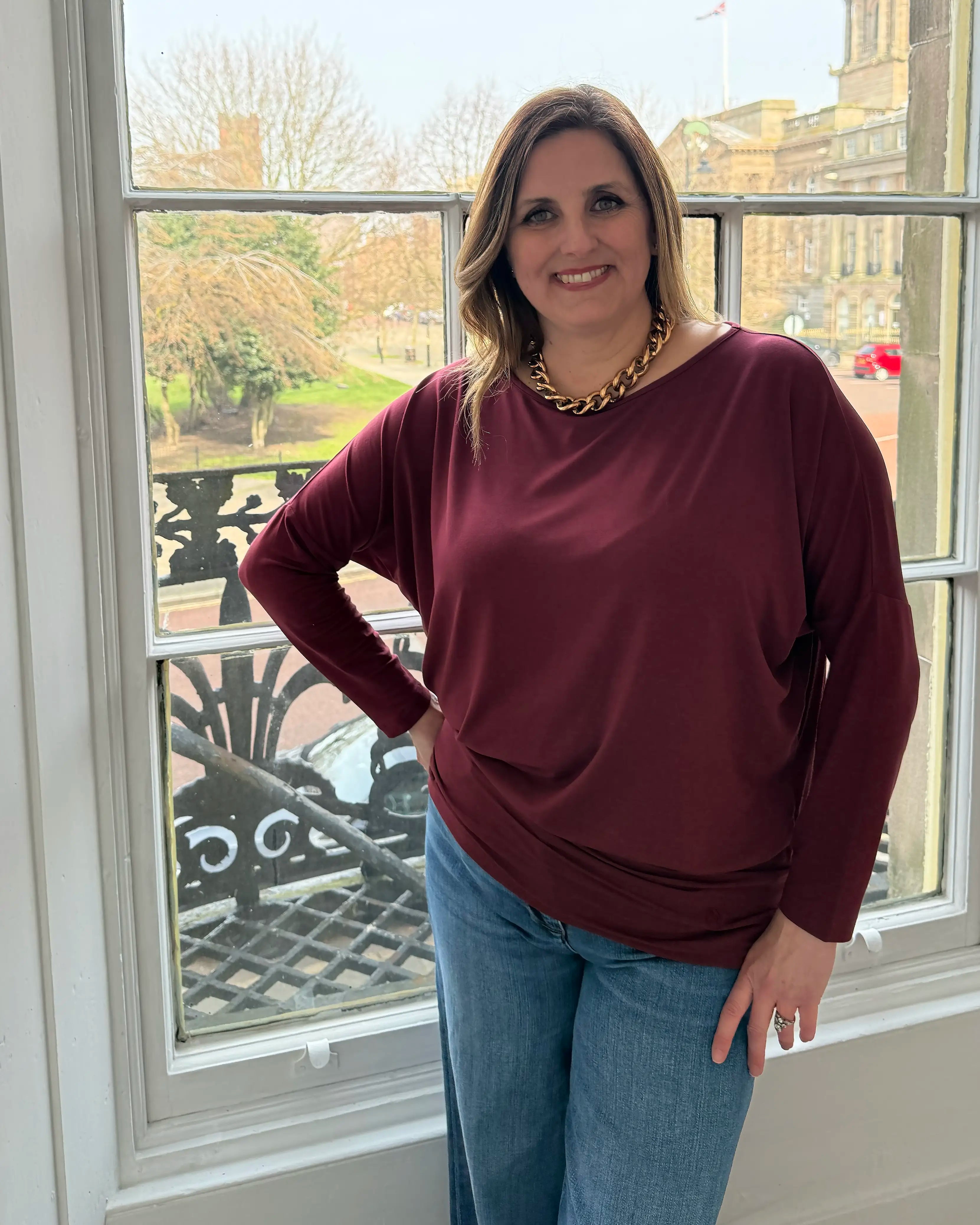Woman wearing a burgundy sweater and blue jeans standing in front of a window with a garden view.