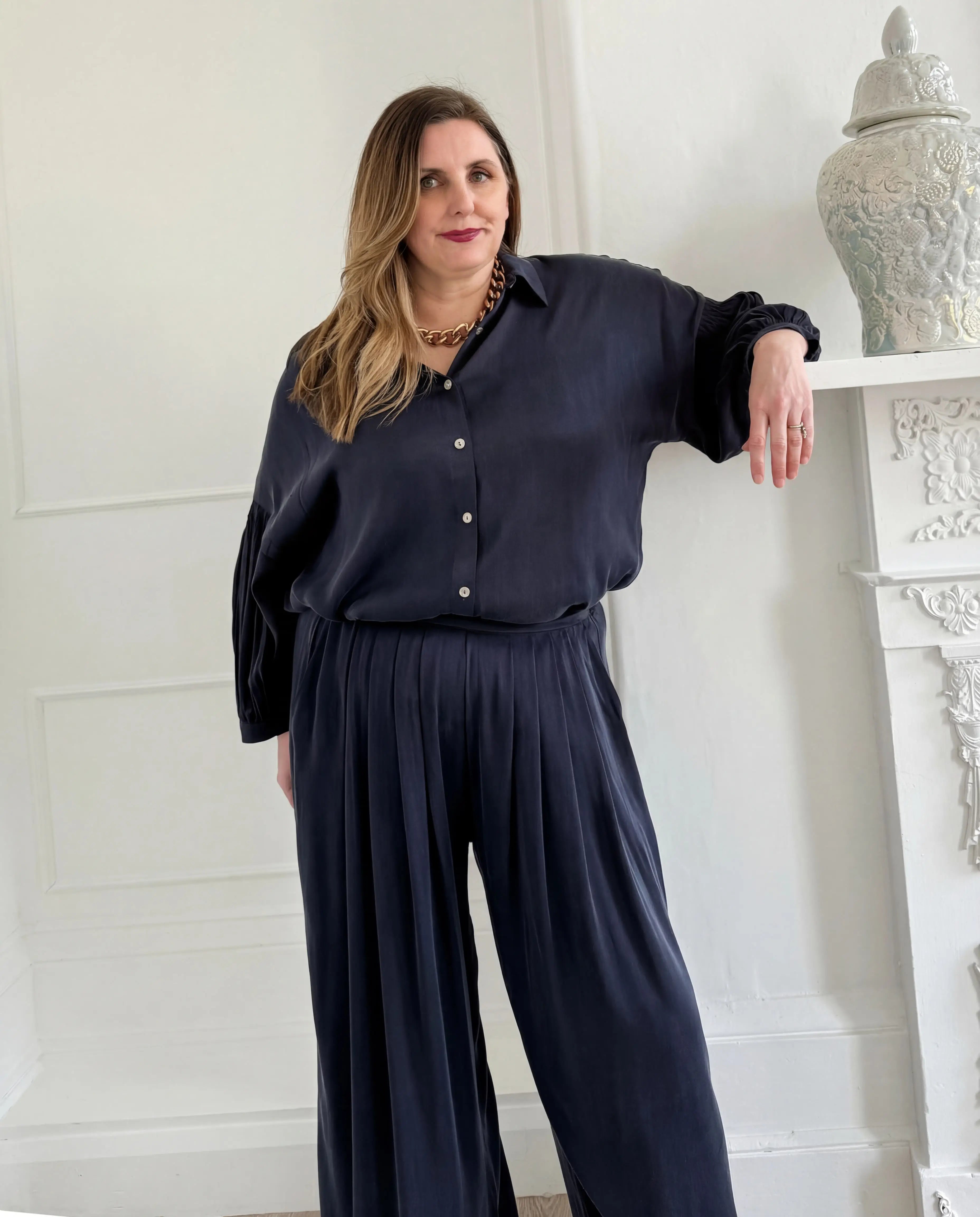Woman wearing a navy Melyr silky navy trouser and shirt standing against a white wall.