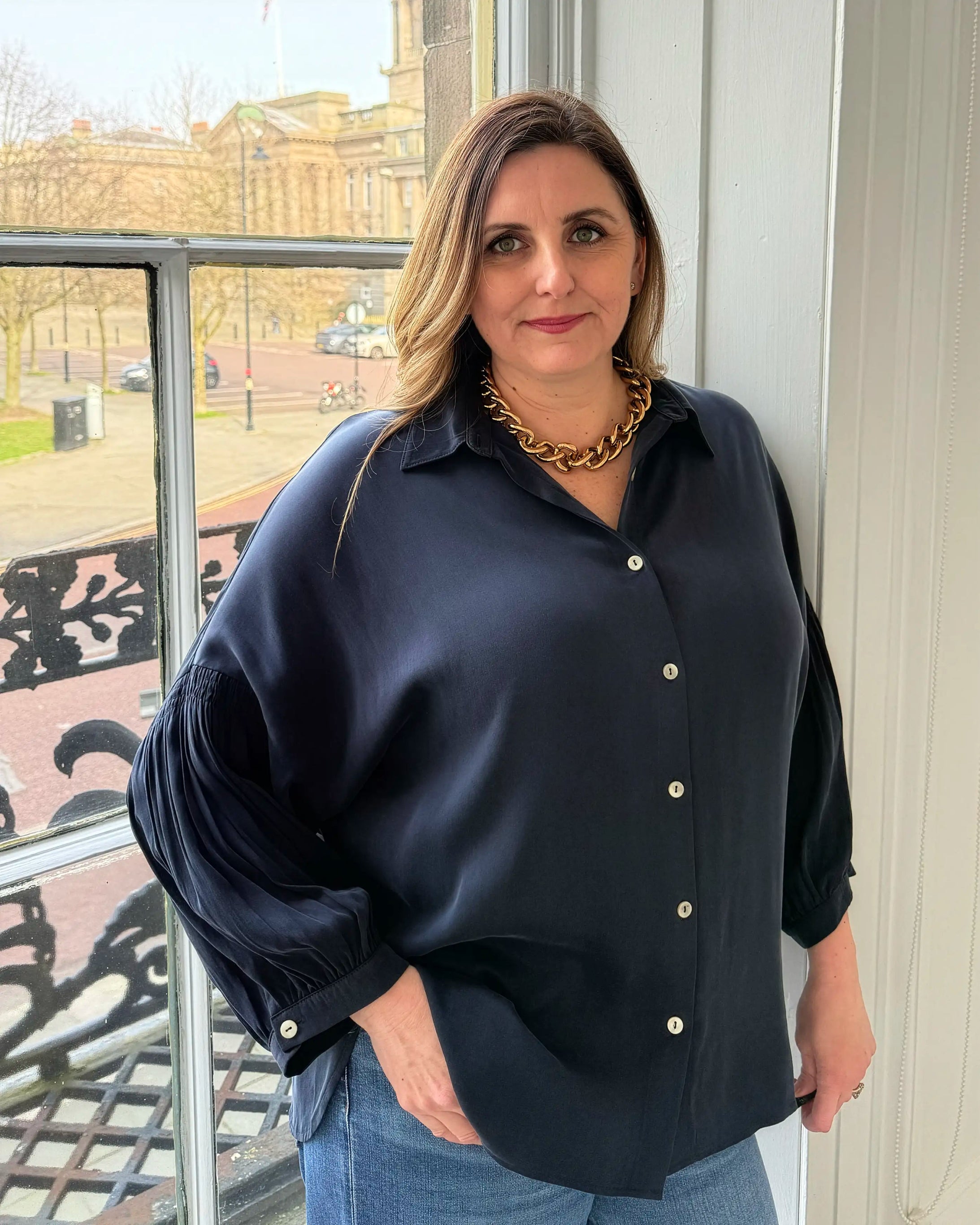 Woman wearing a black blouse with a gold necklace standing indoors.