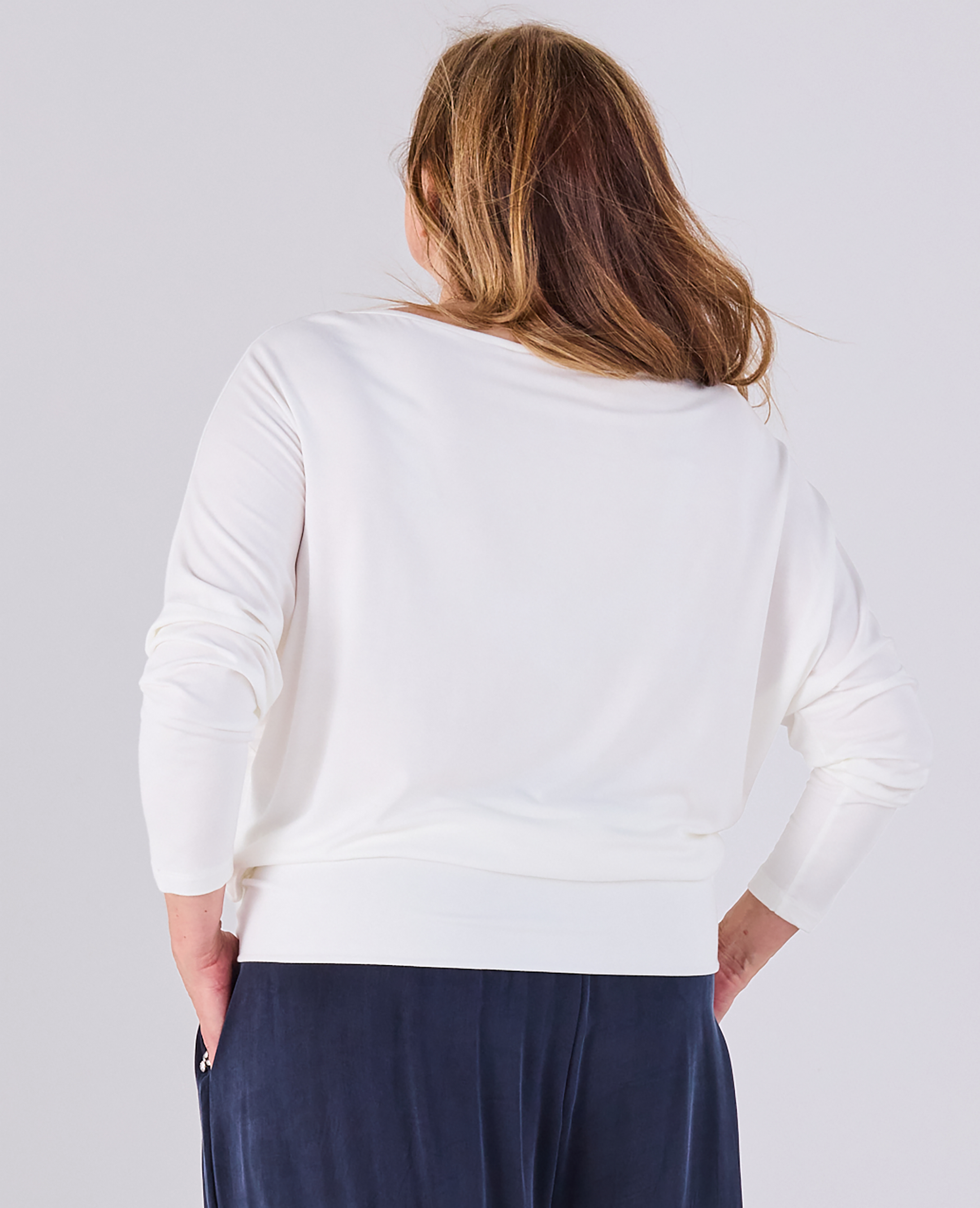 A woman viewed from behind wearing a Melyr Aimee white long-sleeve top and navy blue pants against a light gray studio background.