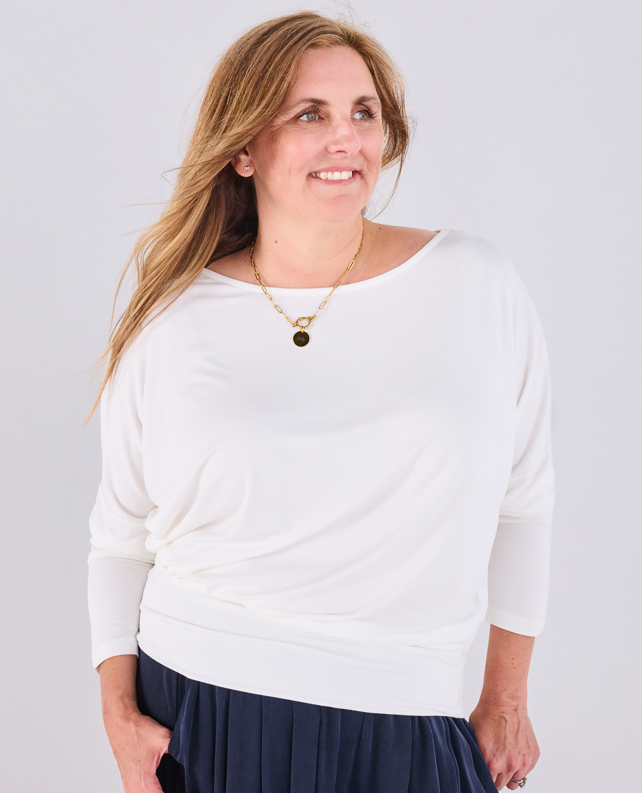 A woman wearing a Melyr white Aimee three-quarter sleeve top with a boat neckline and a navy portofino trousers accessorized with a gold necklace, against a light grey background.