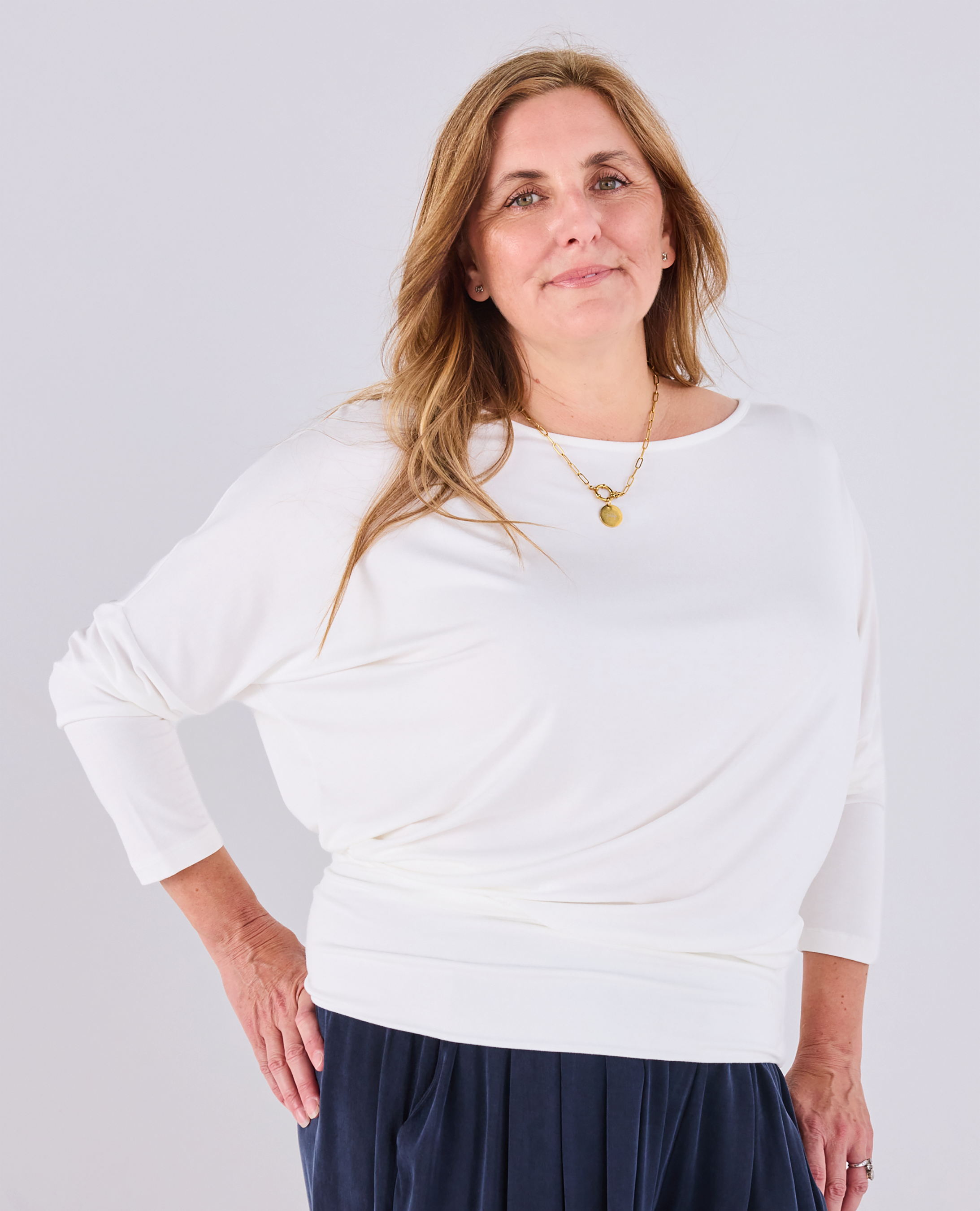 A woman modeling a Melyr white Aimee long-sleeve top with a draped, boat-neck design, paired with navy Portofino pants, against a light grey studio background.