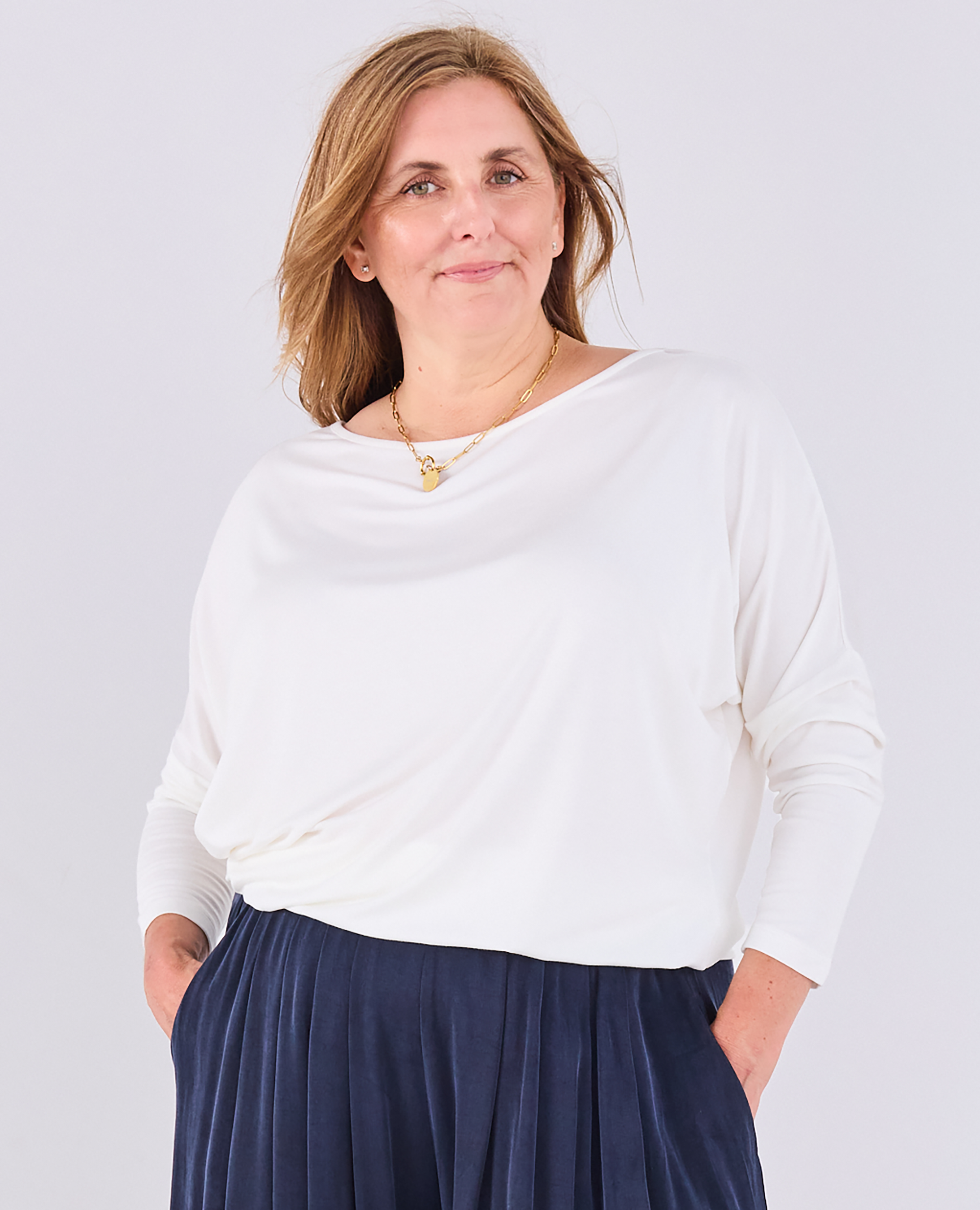 A woman wearing a Melyr white Aimee long-sleeve draped top and navy portofino pleated trousers standing against a light gray studio background.