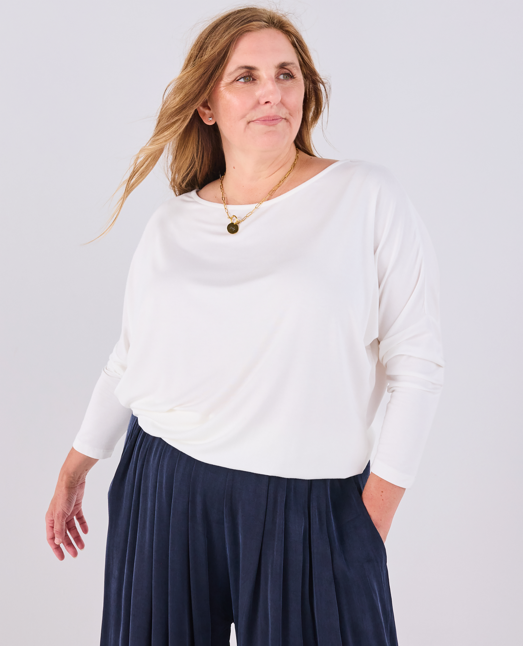 A woman wearing a Melyr white Aimee long-sleeve boat-neck top and navy portofino blue pleated pants, standing against a light grey studio background.