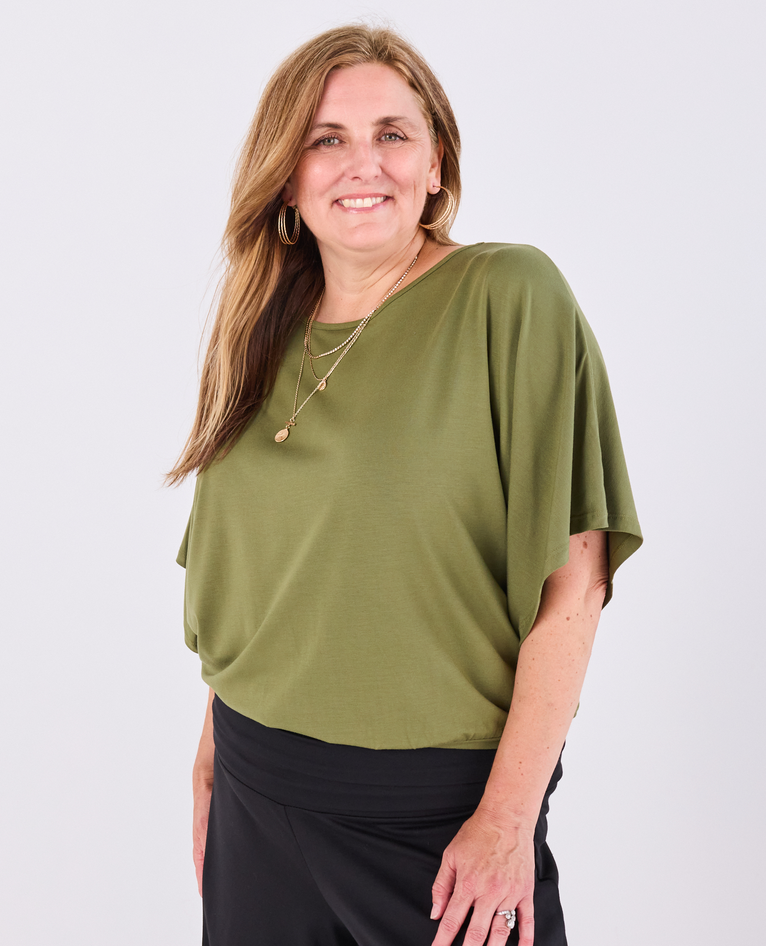 A woman wearing a Melyr, short-sleeve olive green Nico top and black bottoms, standing against a light grey studio background.